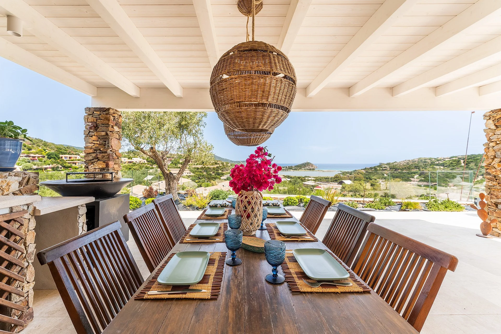 Tavolo da pranzo all'aperto con sei piatti e sei bicchieri blu, decorato con un vaso di fiori rosa e una tovaglia di paglia, sotto un lampadario di vimini, con vista sul paesaggio collinare e il mare.