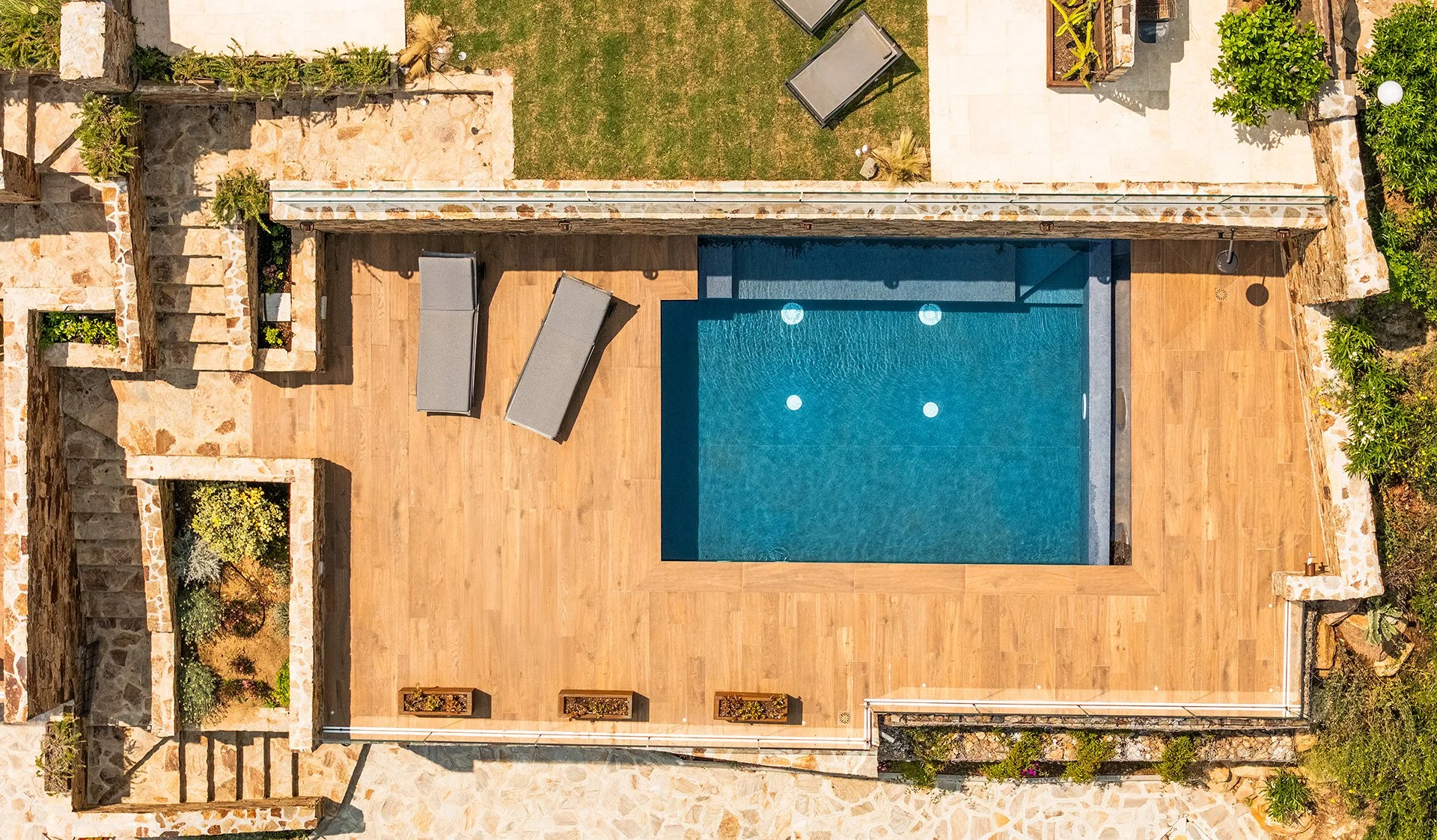 Veduta aerea di una piscina rettangolare con giochi d'acqua, circondata da un pavimento in legno e aree con piante e sedie a sdraio.