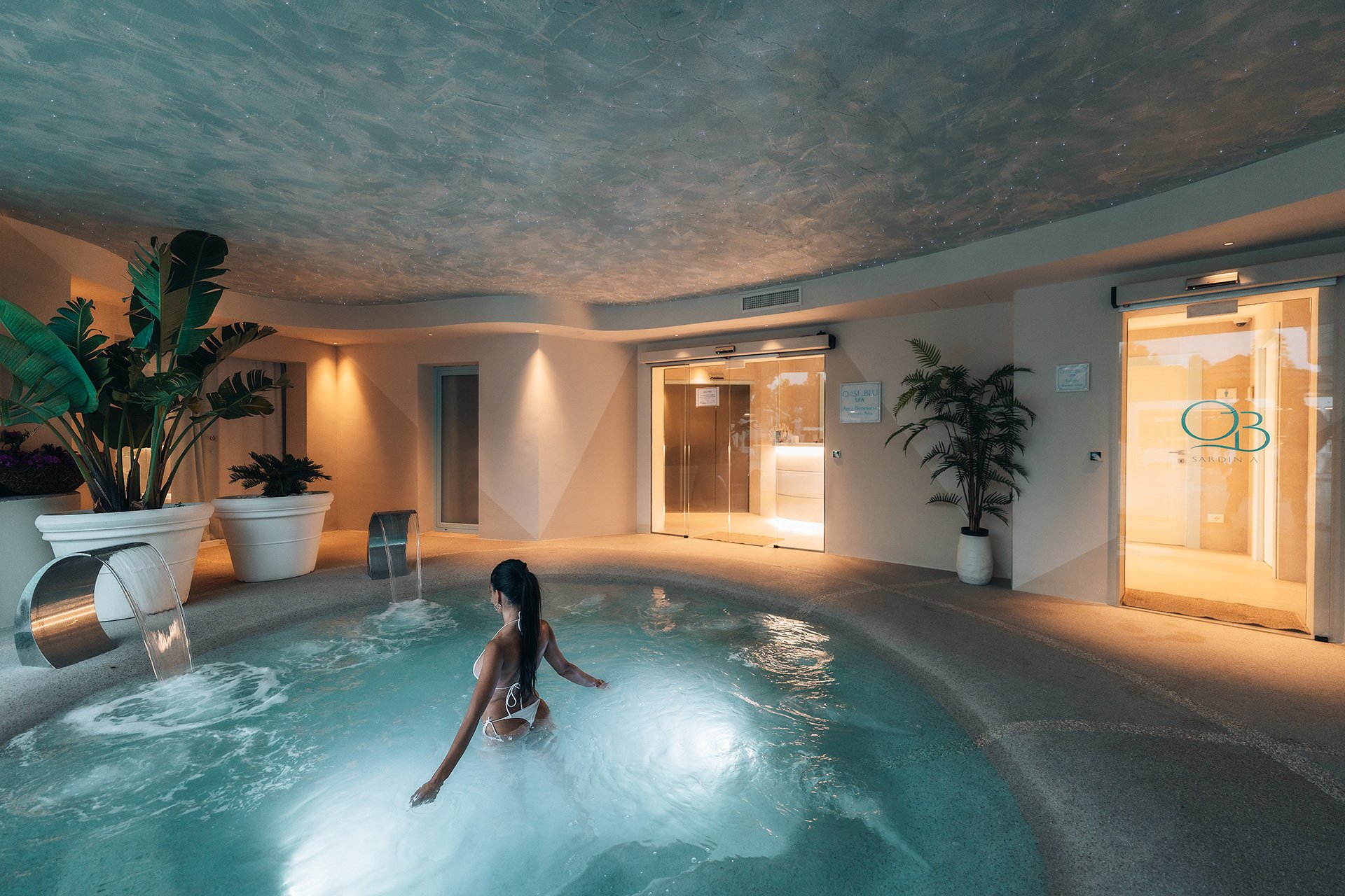 Interno di una spa con una piscina riscaldata, piante decorative in grandi vasi e un soffitto dipinto che imita il cielo stellato. Una donna si rilassa nell'acqua vicino al bordo della piscina, mentre sullo sfondo si vede una porta in vetro con il lo