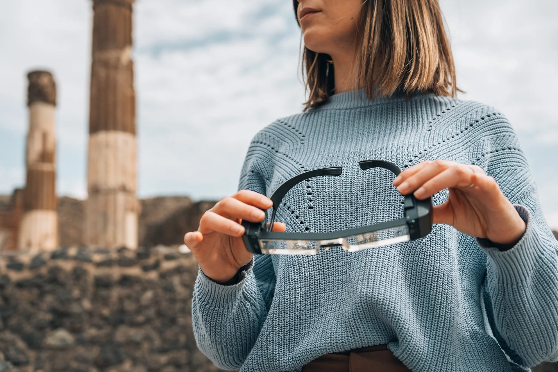 Donna che indossa un maglione azzurro e tiene un visore per realtà virtuale in mano, con un paesaggio di rovine in background.