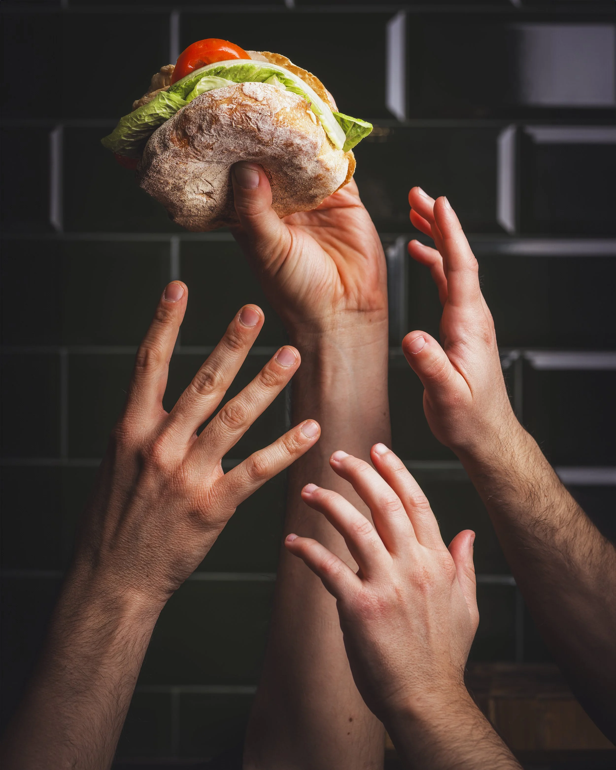 Immagine di più mani che raggiungono un sandwich con pane croccante, lattuga, pomodoro e carne, contro uno sfondo scuro.