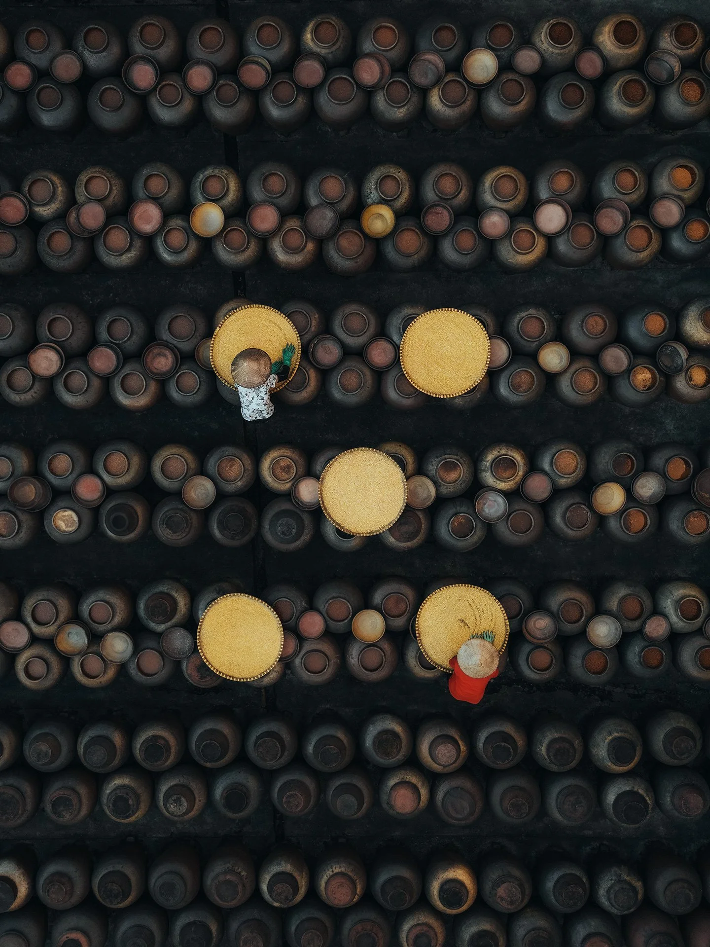 Immagine dall'alto di scaffali con bottiglie di vetro sovrapposte, alcune con il collo rivolto verso l'alto. Ci sono due persone con cappelli di paglia e abiti colorati che sembrano maneggiare o ispezionare le bottiglie.