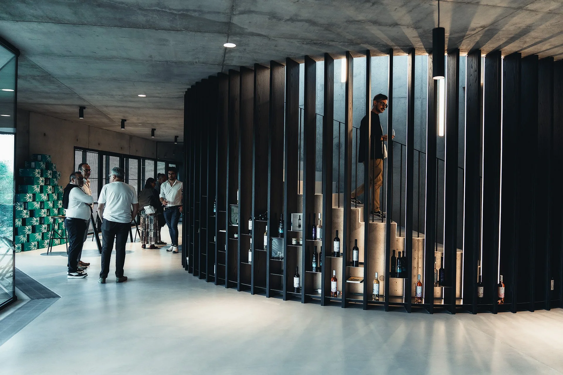 Interno di un edificio moderno con pareti di mattoni, soffitto in cemento e pavimento in cemento. A sinistra, un gruppo di persone sta conversando, con alcuni davanti a uno stand di decorazioni verdi. Al centro, una struttura a scaffale con bottiglie di vino è posizionata vicino a una scala di pietra, con una persona in piedi su di essa. La scena è illuminata da luci a soffitto e da luci verticali lungo le pareti.