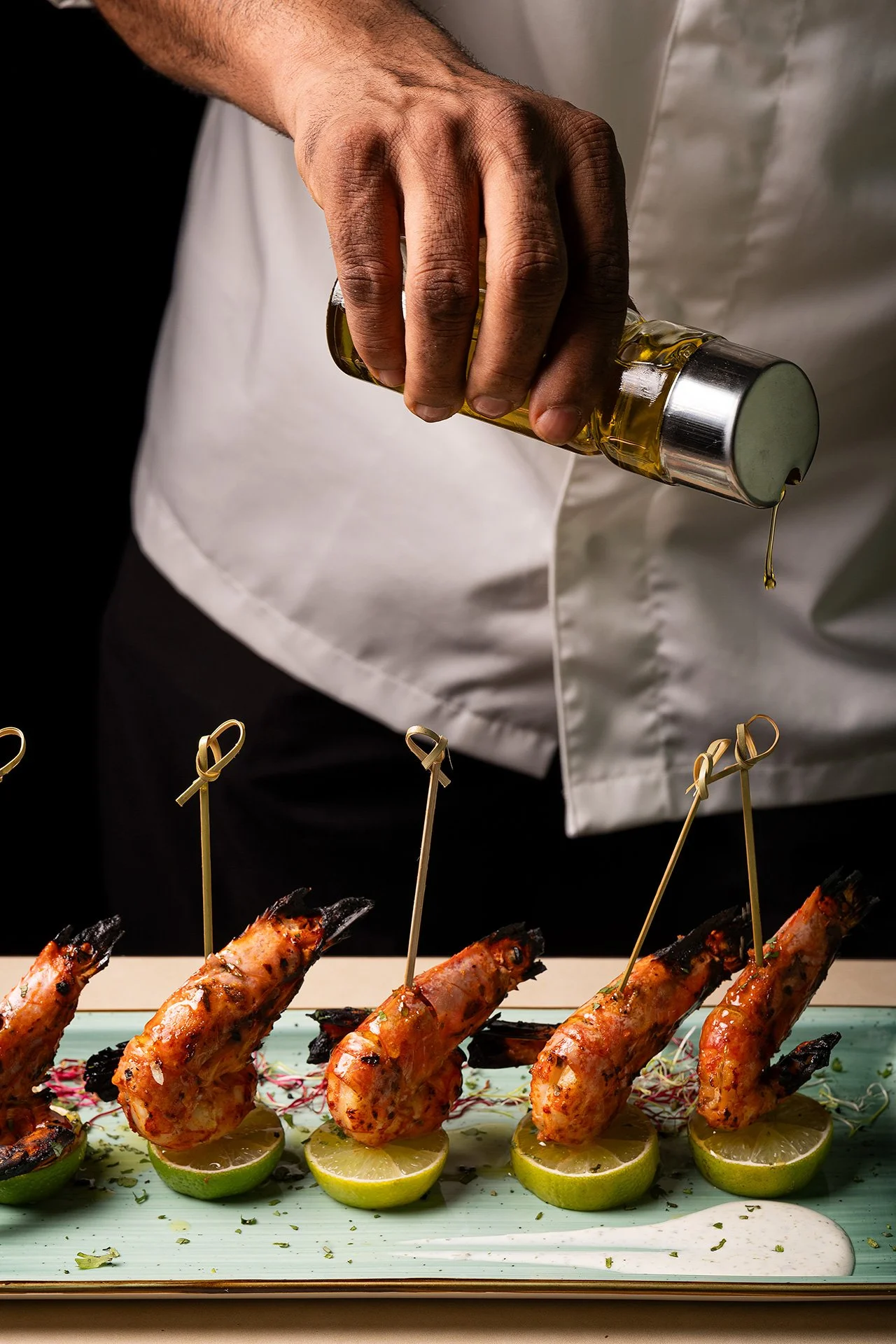 Una mano che versa olio su gamberi grigliati posizionati su fettine di lime, decorati con erbe e spezie, serviti su un piatto con una salsa bianca accanto.