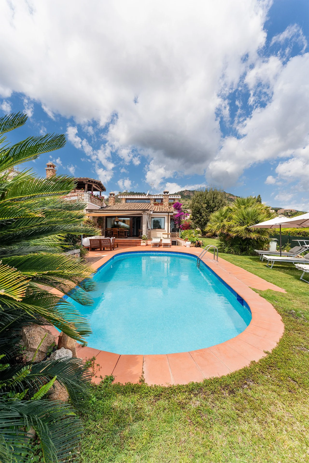 Piscina all'aperto in un giardino con lettini, ombrelloni, piante tropicali e una casa sullo sfondo sotto un cielo nuvoloso.