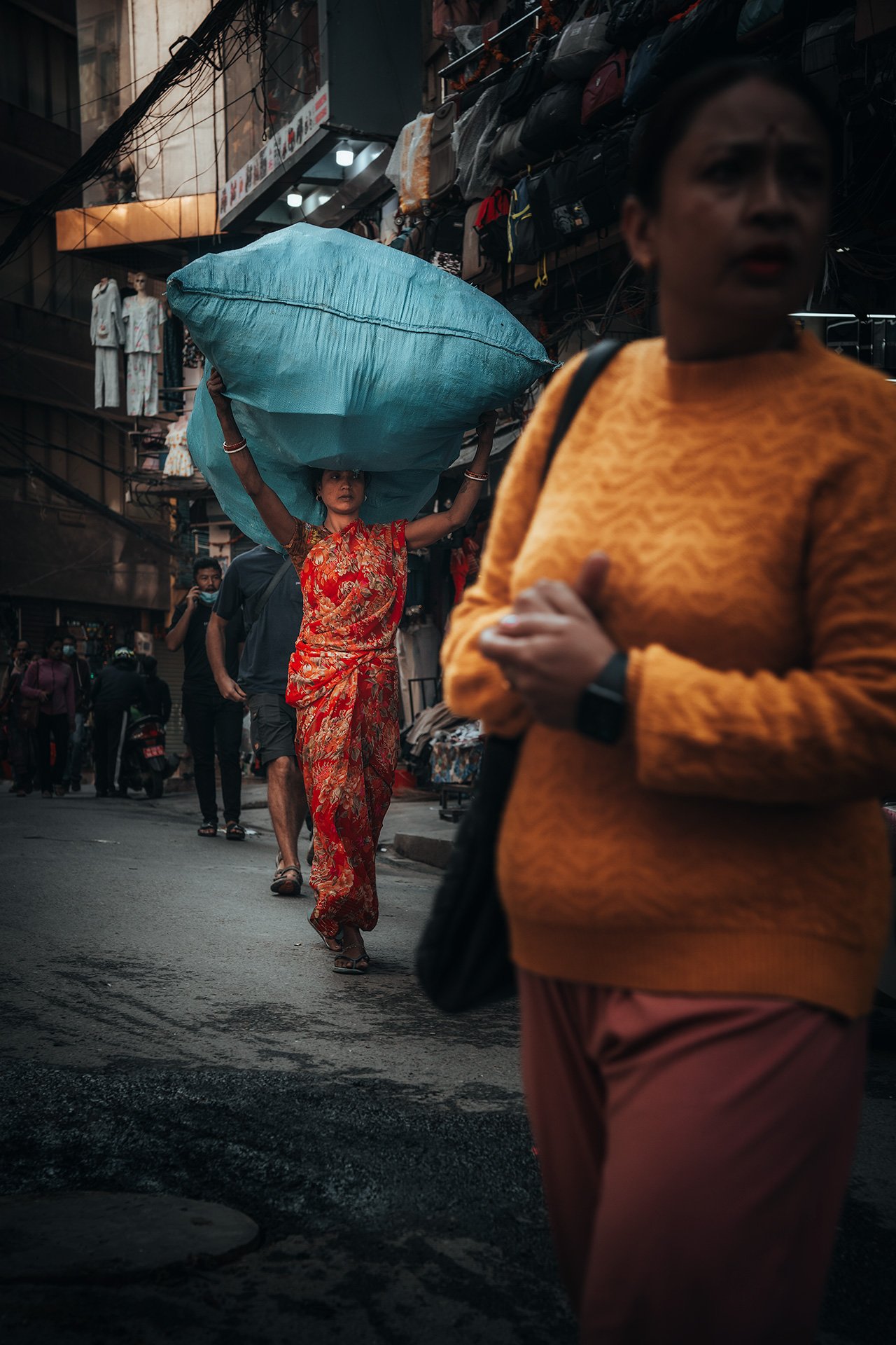 Una donna indossa un sari rosso mentre cammina per strada mentre una donna con una maglia gialla passa di fronte a lei, in un mercato affollato con negozi di abbigliamento e persone che camminano. La scena sembra essere ambientata in un'area urbana a