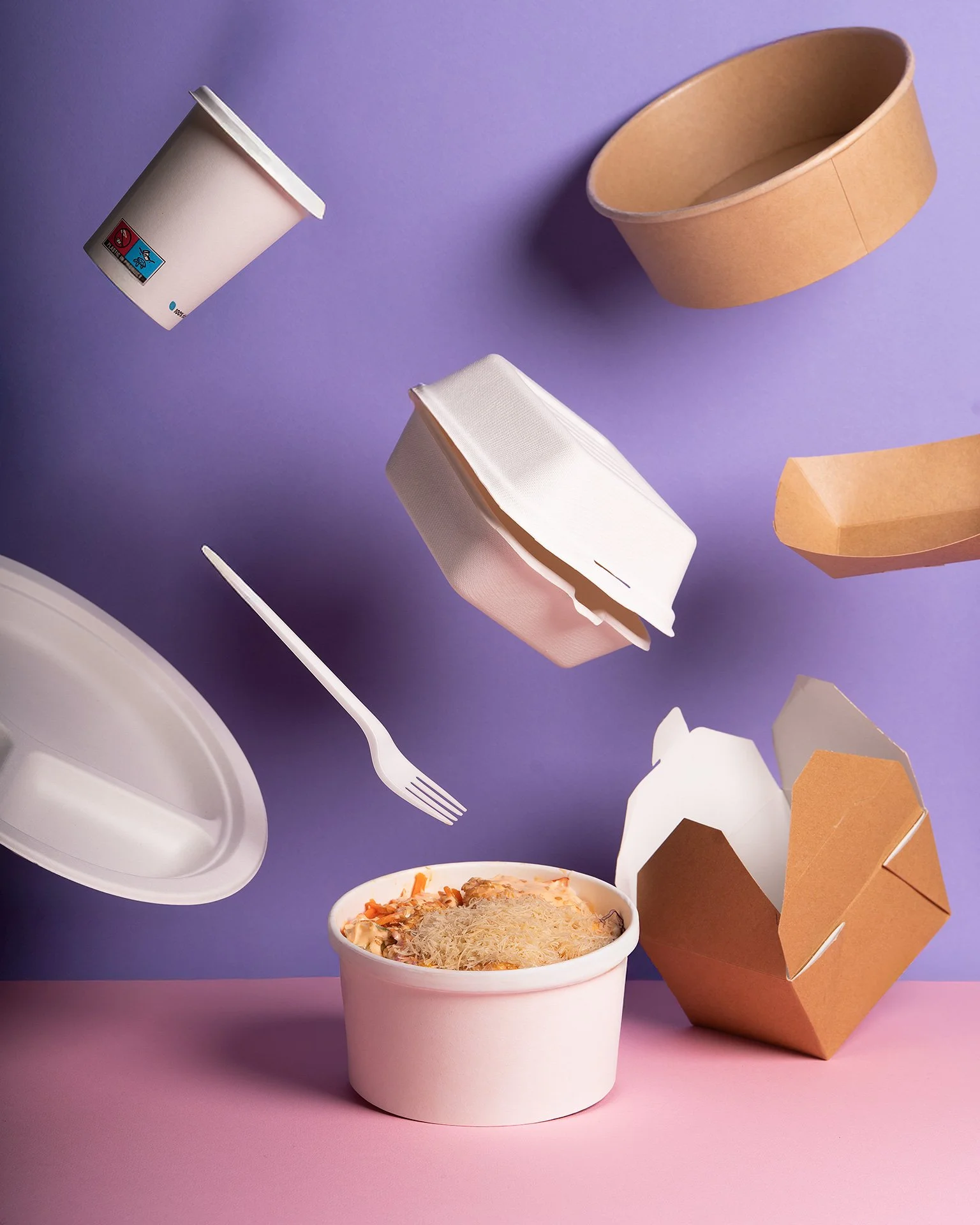 Contenitore di cibo con pasta, torte e divani di cartone, bicchieri e posate, su sfondo viola e rosa.
