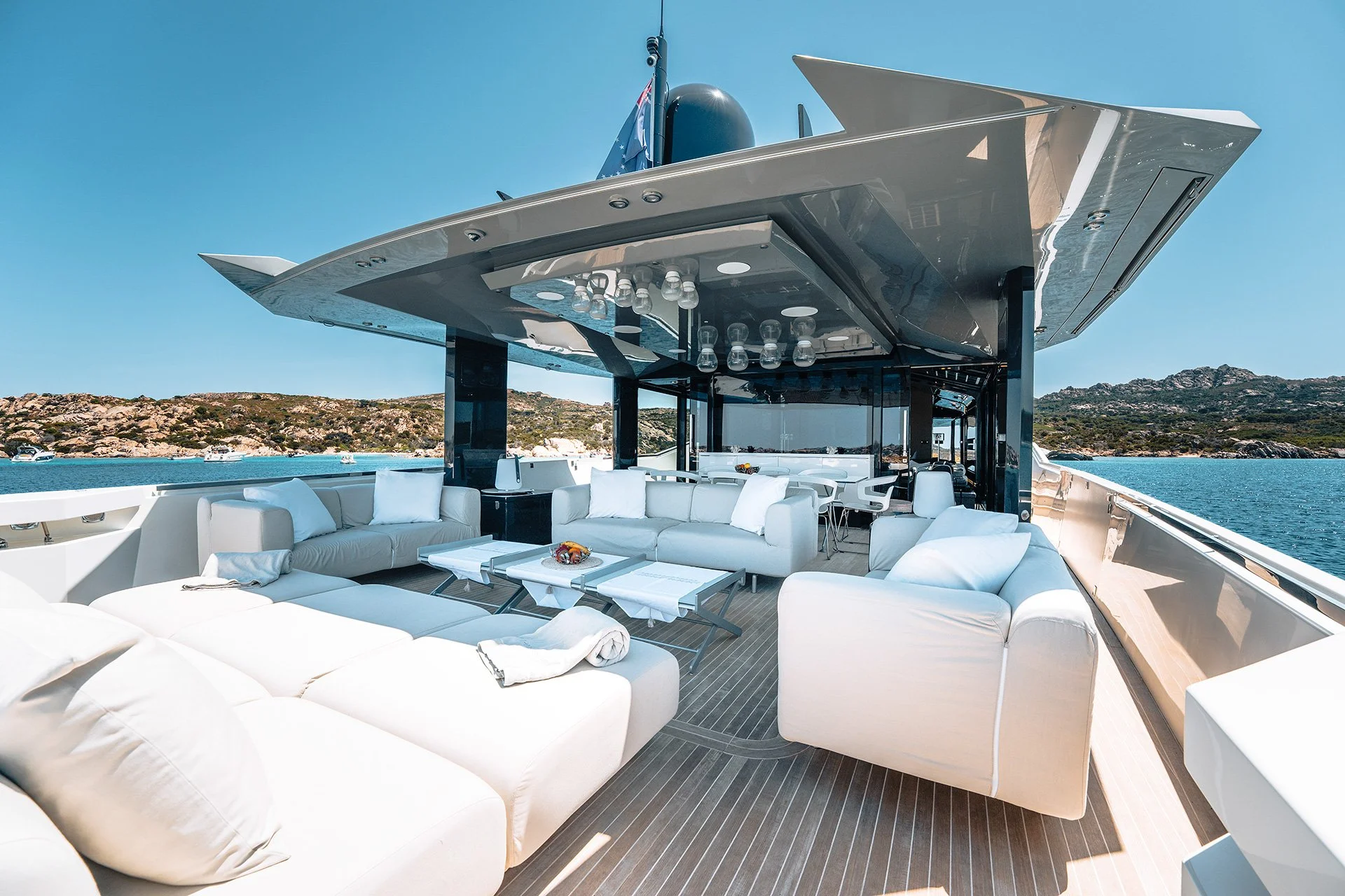 Zona lounge di un yacht con divani bianchi, teli e tavoli, vista mare e isole sullo sfondo, sovrastata da una sovrastruttura con luci e decorazioni moderne.