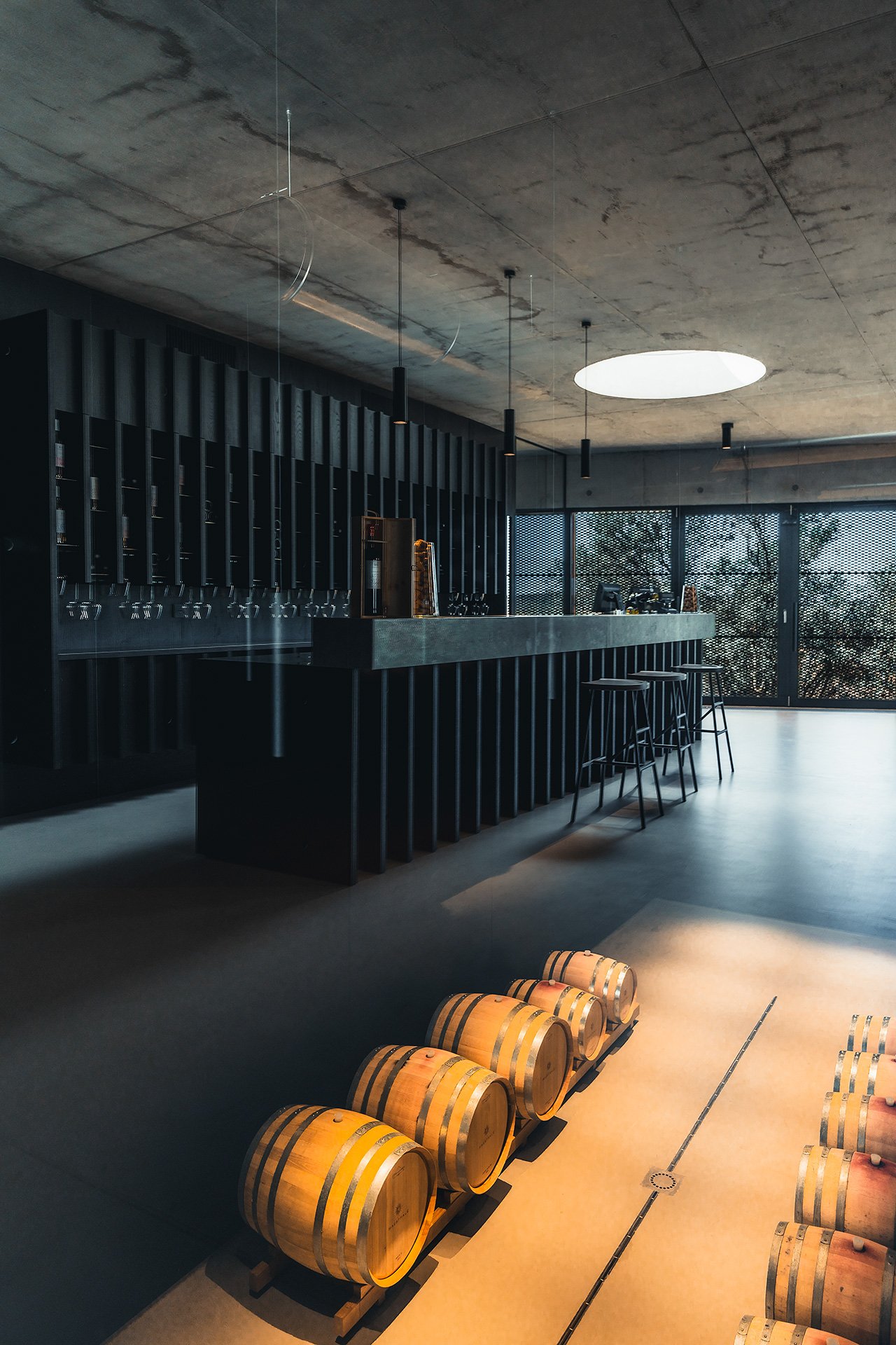 Interno di una cantina moderna con barili di legno in primo piano, un bancone di bar con sedie dietro, illuminazione minimalista e grandi finestre con vista sugli alberi che lasciano entrare la luce naturale.