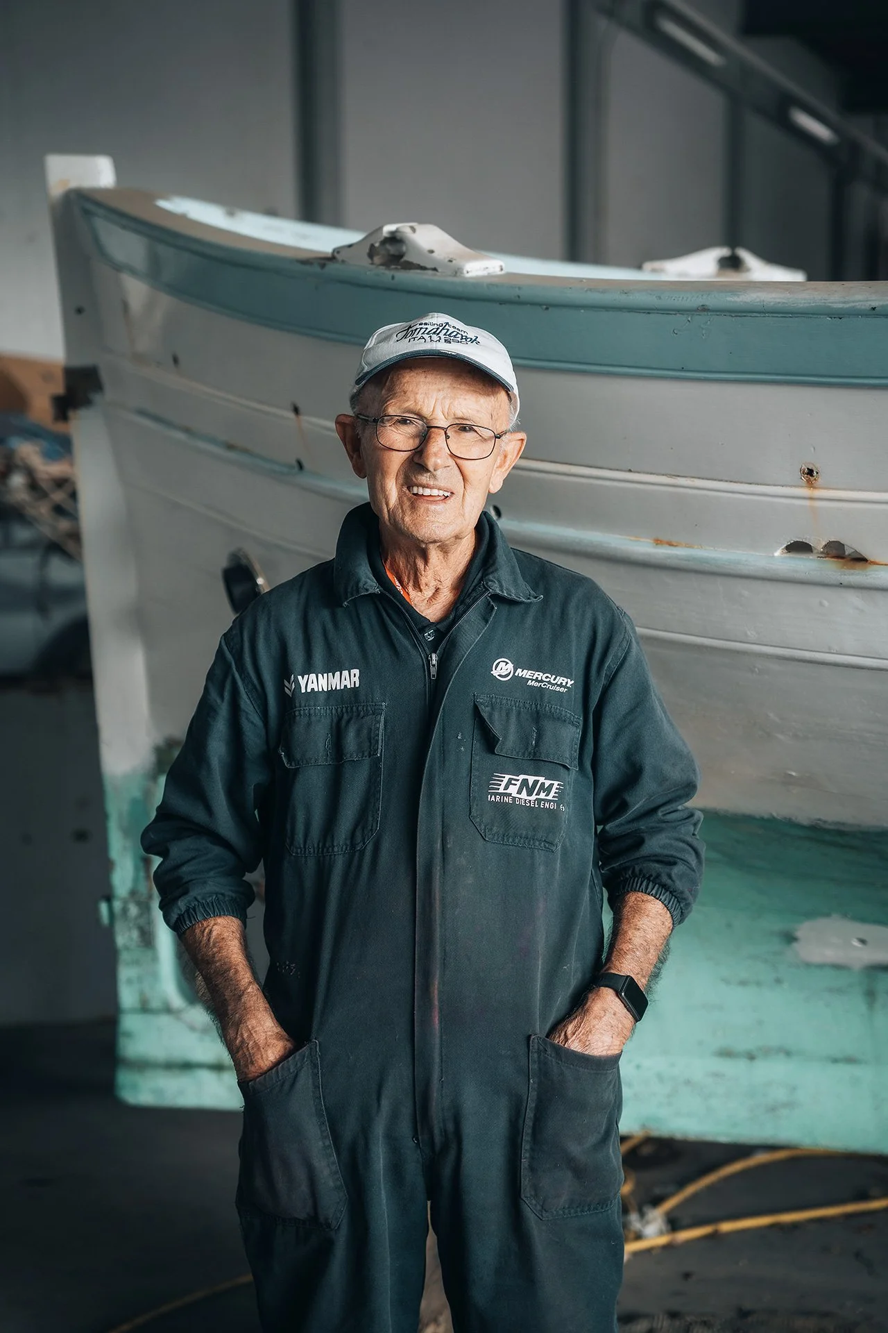 Uomo anziano che indossa una giacca con loghi di aziende di nautica, cappellino e occhiali, in piedi davanti a una barca in un capannone