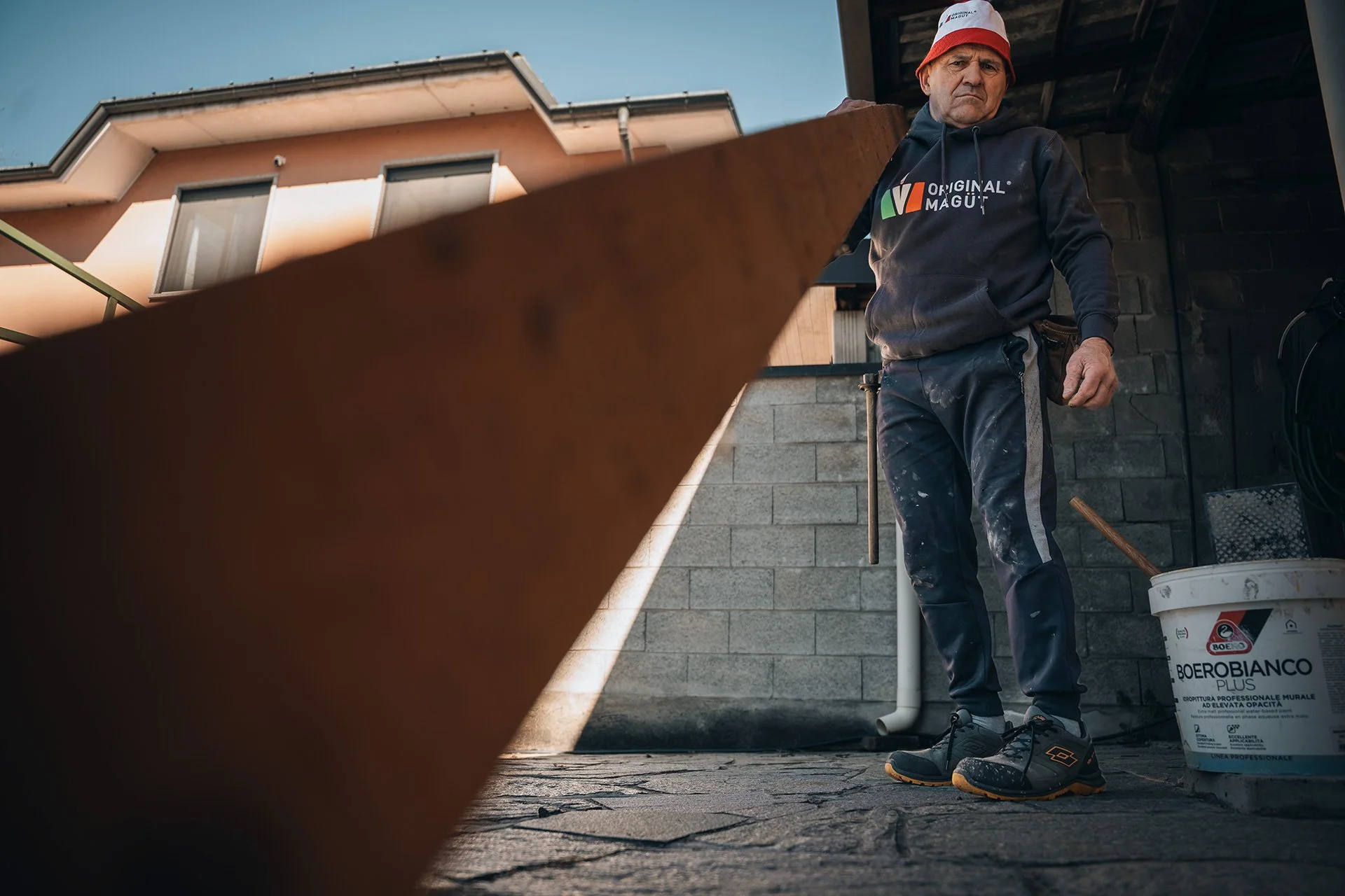 Uomo con cappello calpestato e felpa con logo 'Original Mug' che lavora su un progetto di ristrutturazione di un edificio, con secchio di pittura di fronte e una parete di mattoni sullo sfondo.