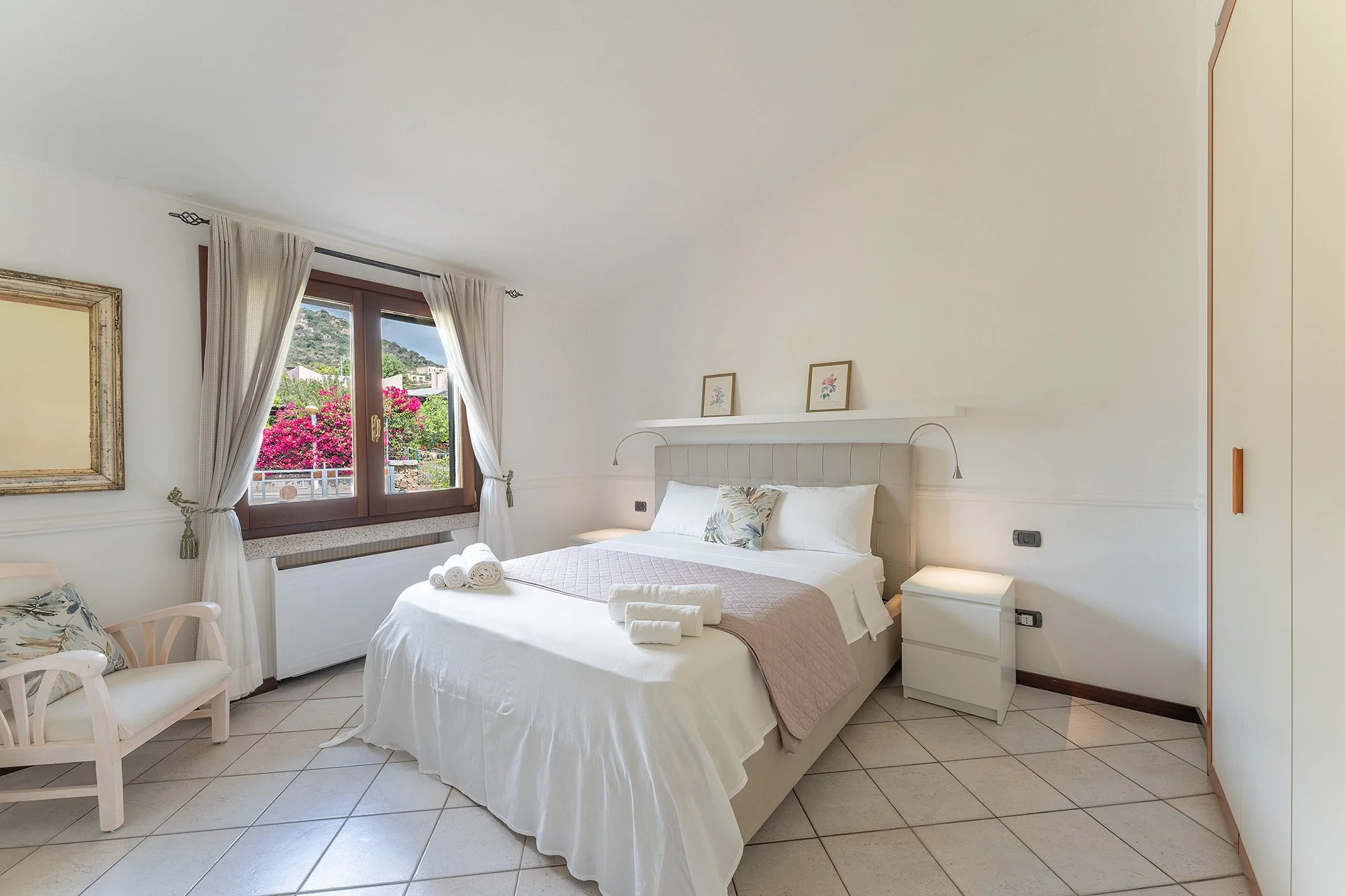 Camera da letto luminosa con letto matrimoniale, vista sulla finestra con tende bianche e paesaggio verde, decorazioni floreali e quadri alle pareti, mobili bianchi e c'è un'armadio a muro.