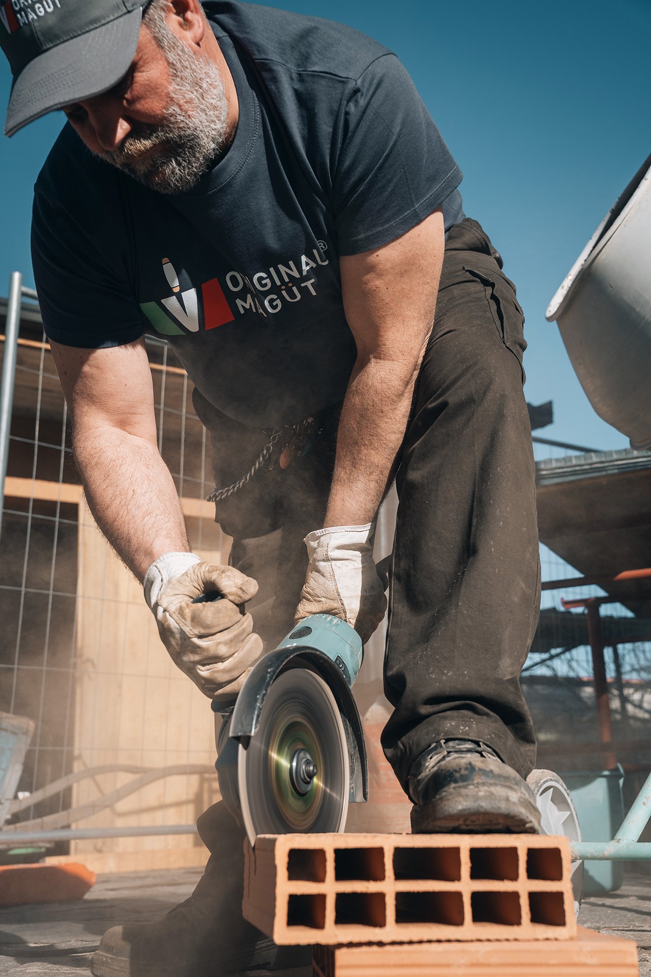 Uomo che usa una smerigliatrice su un mattone di laterizio, indossando guanti e abbigliamento da lavoro, in un cantiere edile all'aperto con struttura in costruzione e cielo azzurro.