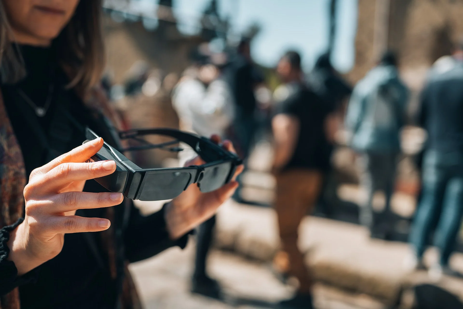 Persona che utilizza uno smartphone con inquadratura ravvicinata delle mani, gruppo di persone in piedi e conversano in un'area pubblica esterna.