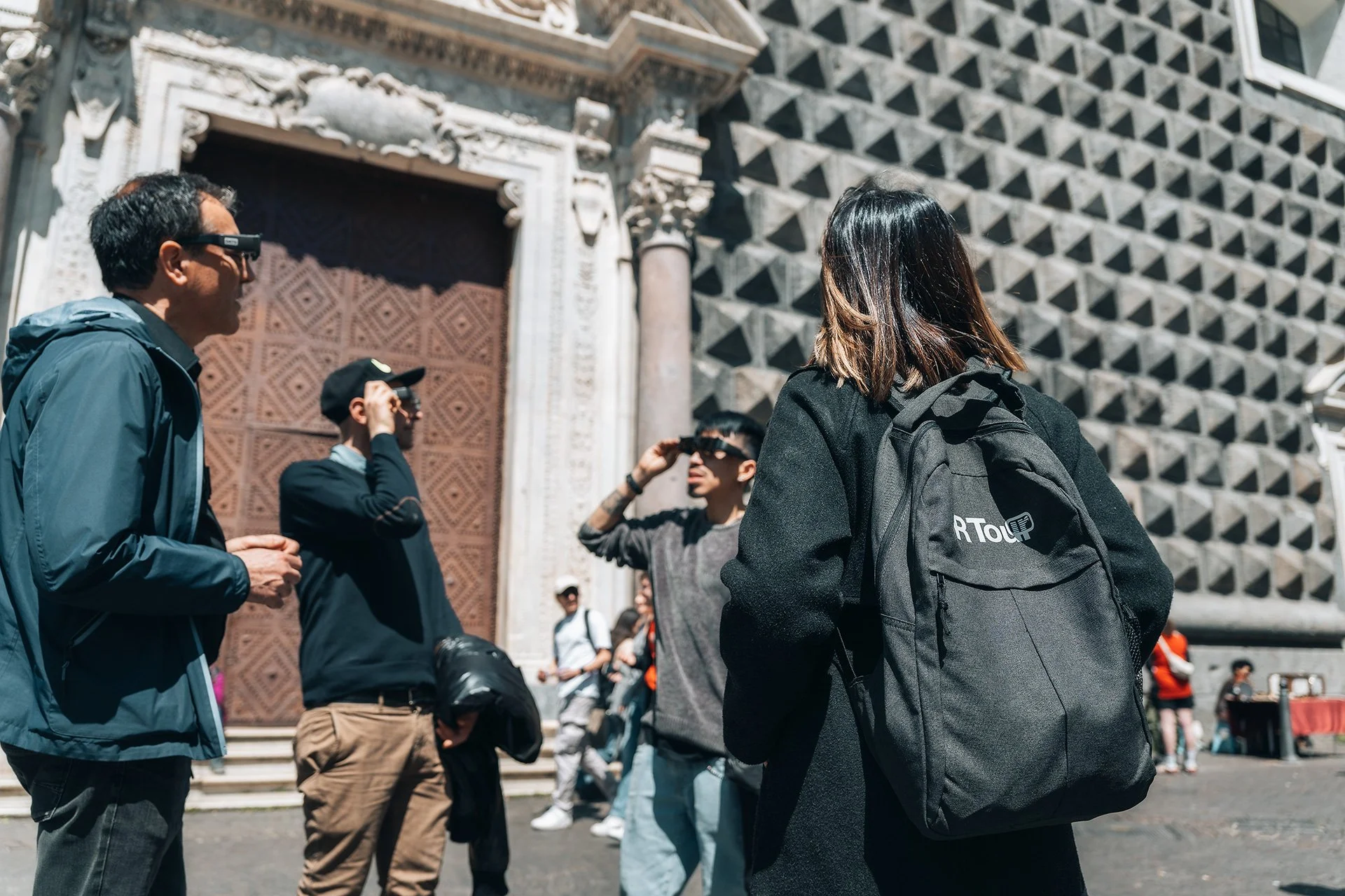 Gruppo di turisti davanti a un edificio storico, con alcuni che indossano occhiali da sole e altri con zaini, mentre parlano e scattano foto.