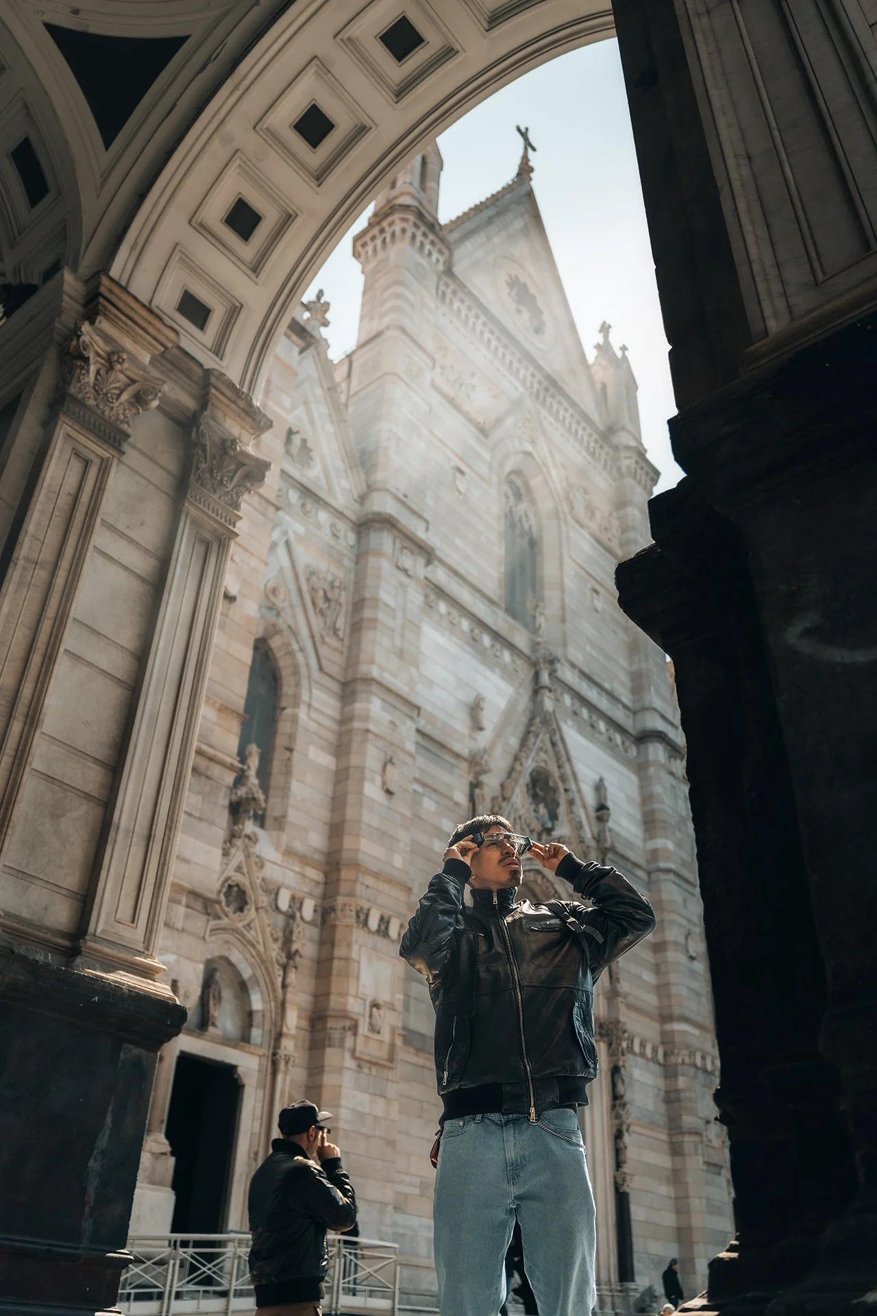 Un uomo in giacca di pelle prende una foto davanti a una cattedrale gotica con archi e torri, durante il giorno.