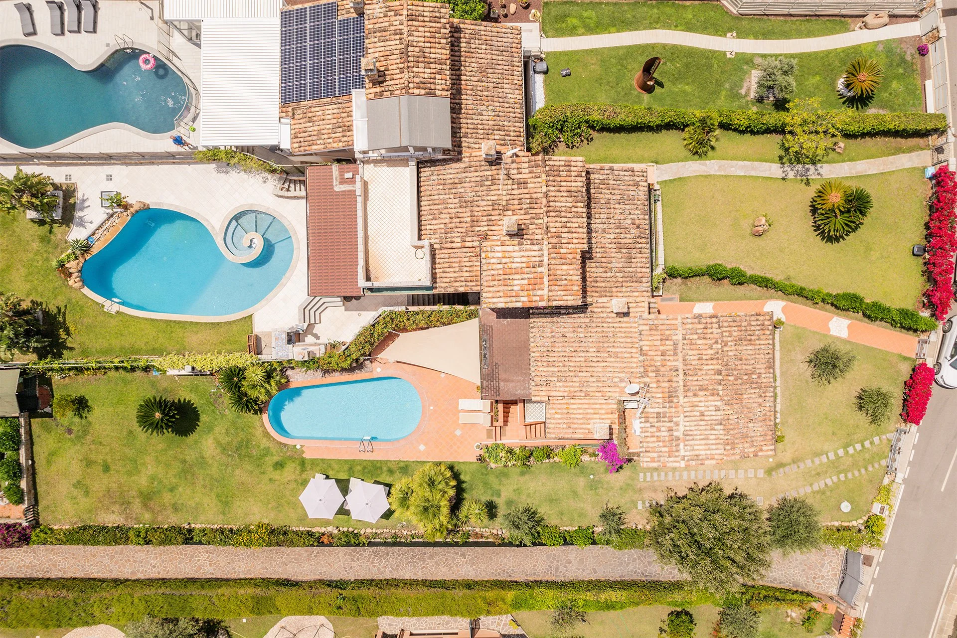 Veduta aerea di un giardino con due piscine, una di forma irregolare e una rettangolare, circondate da piante, alberi e una strada.