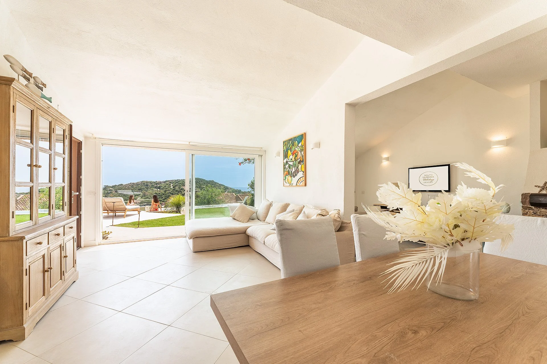 Interno di un soggiorno con divano bianco, pareti bianche e una grande porta scorrevole che apre su un giardino con vista mare e due persone che si godono il paesaggio. Arredi in stile minimalista con una decorazione floreale in primo piano su un tav