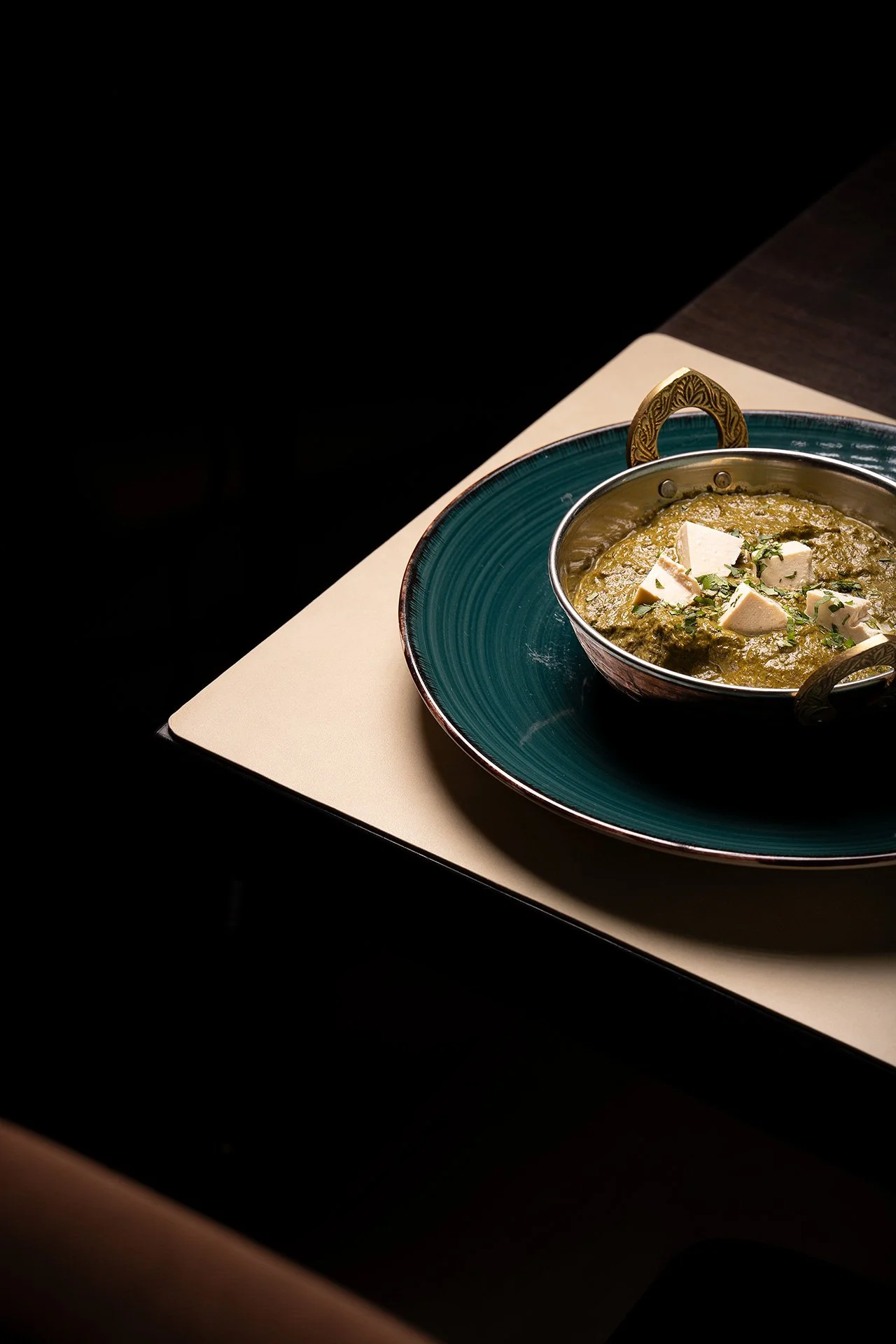 Una ciotola con curry verde, formaggio a cubetti e erbe, appoggiata su un piatto blu scuro, sul tavolo in un ambiente con illuminazione soffusa.