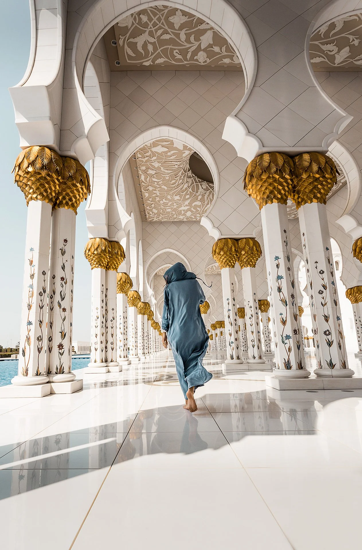 Una persona vestita con abiti tradizionali si passeggia tra colonne decorate con motivi floreali in un'architettura bianca e dorata, con dettagli di foglie dorate sui capitelli, all'aperto sotto un cielo limpido.