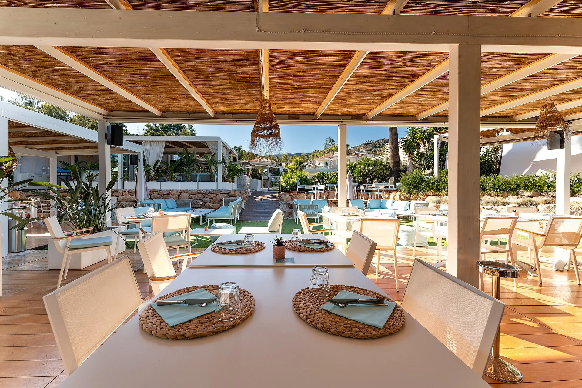 Immagine di un'area all'aperto di un ristorante o bar con tavoli e sedie, un camino o una stufa laterale, con copertura in legno e decorazioni in vimini, piante verdi e vista sul paesaggio sopra le costruzioni.