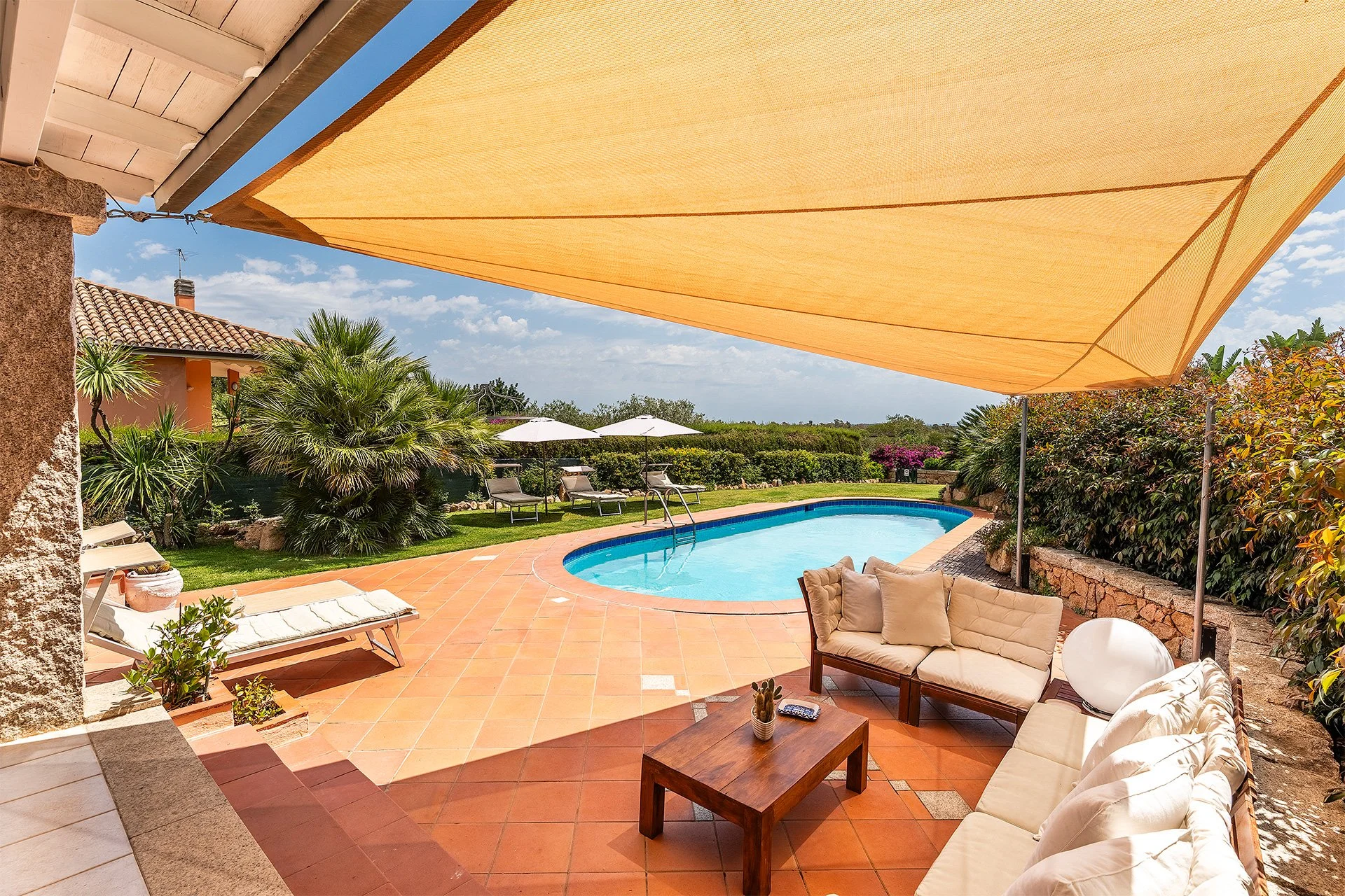 Zona relax con piscina all'aperto, sedie a sdraio e un'ampia area coperta con divanetti, circondata da piante e un giardino ben curato sotto un cielo azzurro con nuvole leggere.