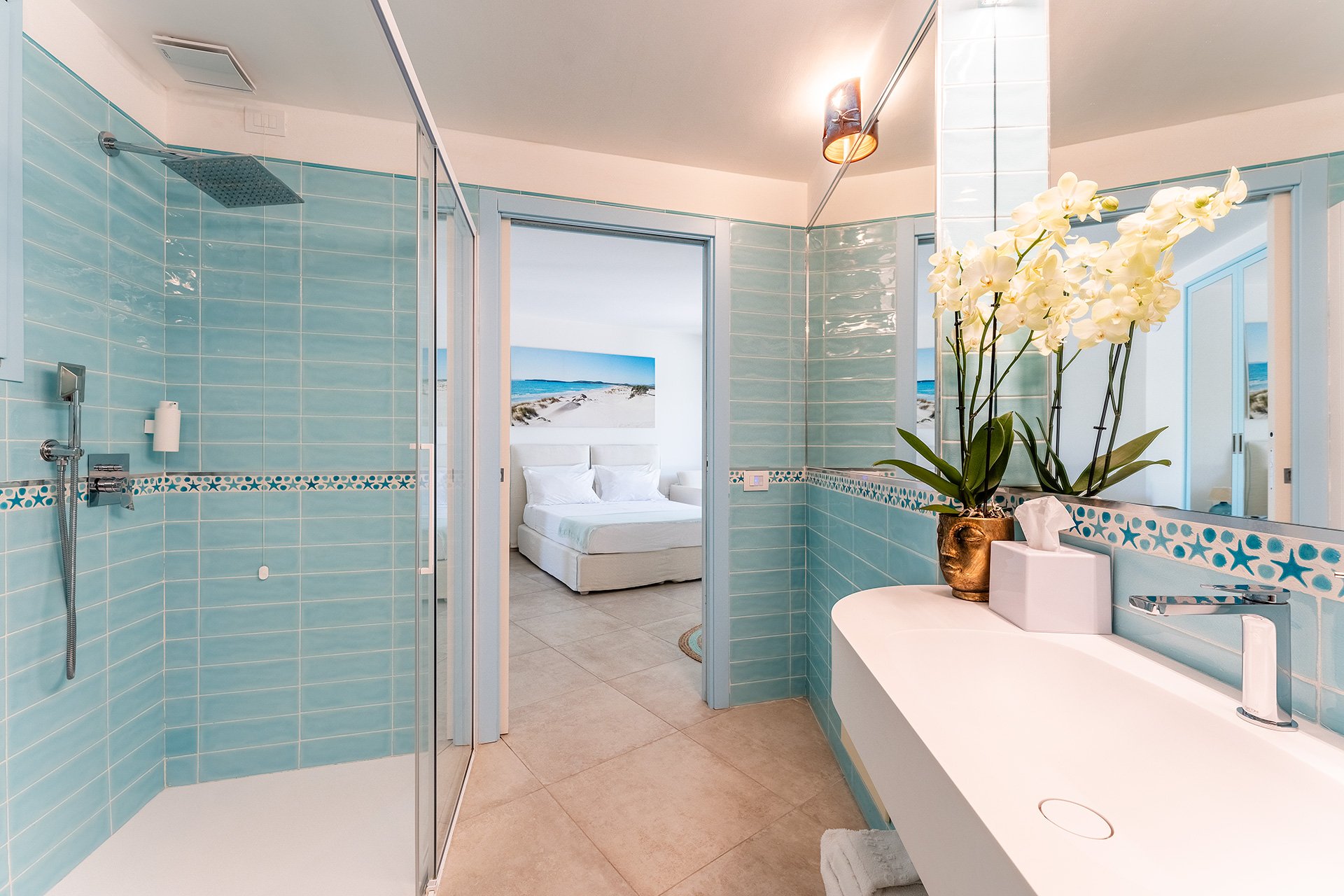 Bagno con pareti azzurre, doccia con pareti di vetro, grande specchio, vaso con orchidee bianche, lavandino sospeso, pareti biene, vista camera da letto con letto bianco, decorazione con quadro di spiaggia, luce calda.