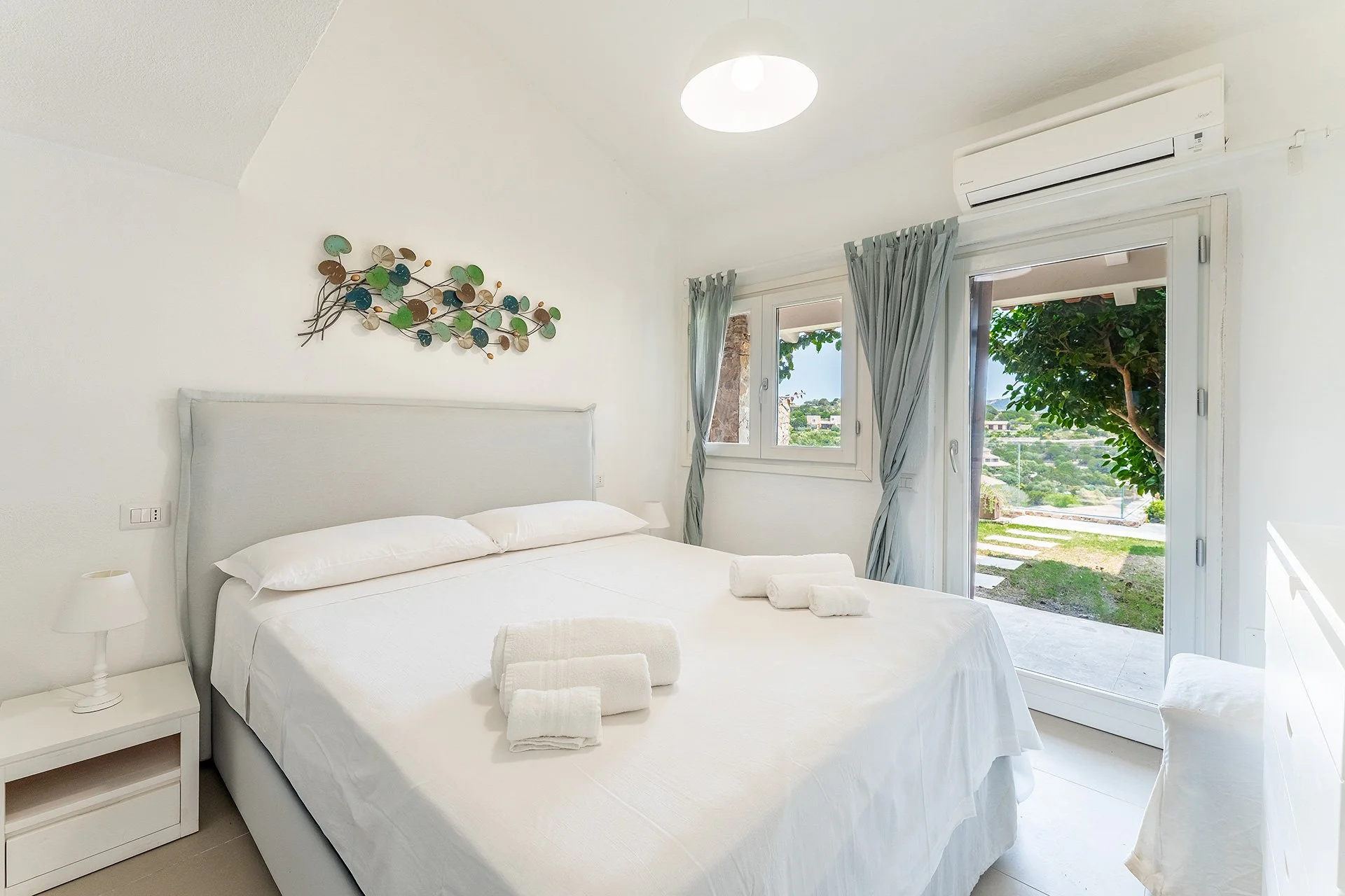 Camera da letto luminosa con letto bianco, due asciugamani piegati sul letto, grande finestra con vista su un giardino verde e porta con tende, aria condizionata a parete, decorazione moderna sulla parete.
