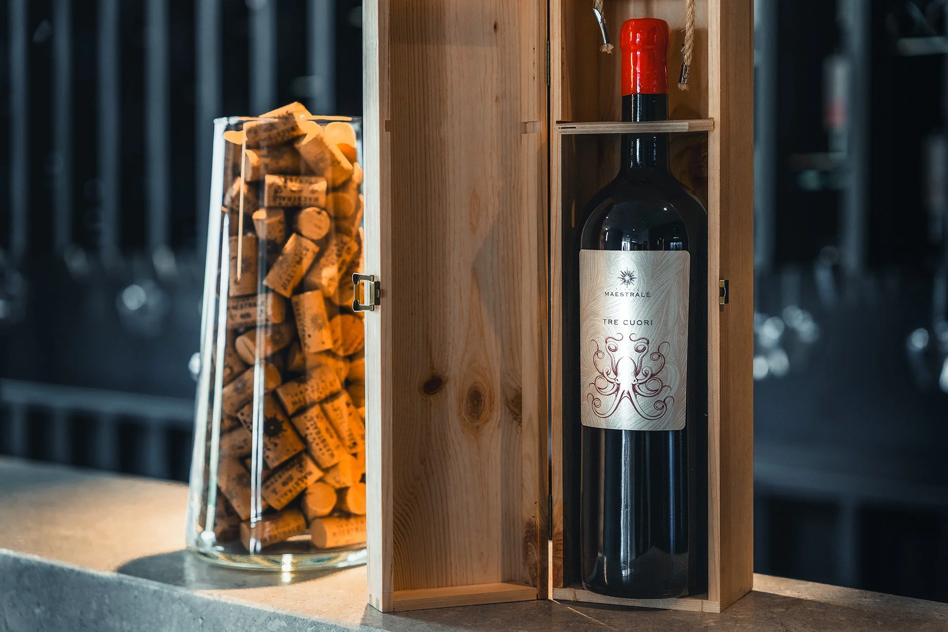 Bottiglia di vino in una scatola di legno, con un tappo rosso, situata tra un contenitore di tappi di sughero e una parete di sfondo con bottiglie di vino.