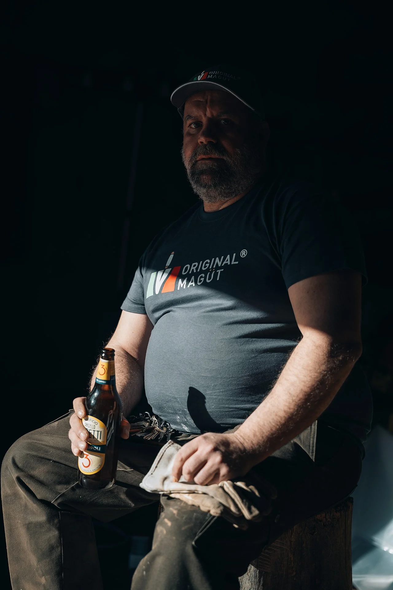 Uomo con barba, cappellino e maglietta grigia seduto in controluce che tiene in mano una bottiglia di birra e un panno
