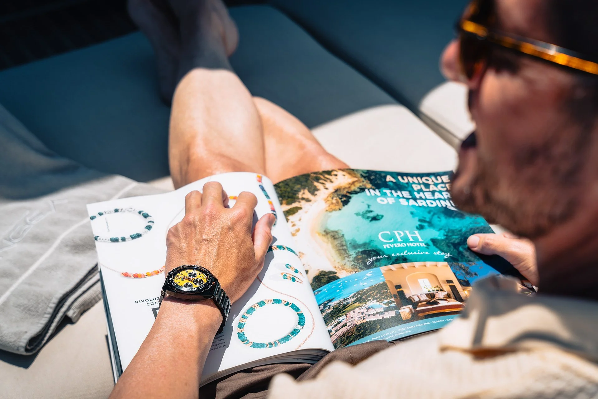 Persona che legge una rivista con immagini di gioielli e un catalogo su un tavolo, con un braccialetto di perline in mano e una vista di una costa in Sardegna nel catalogo.