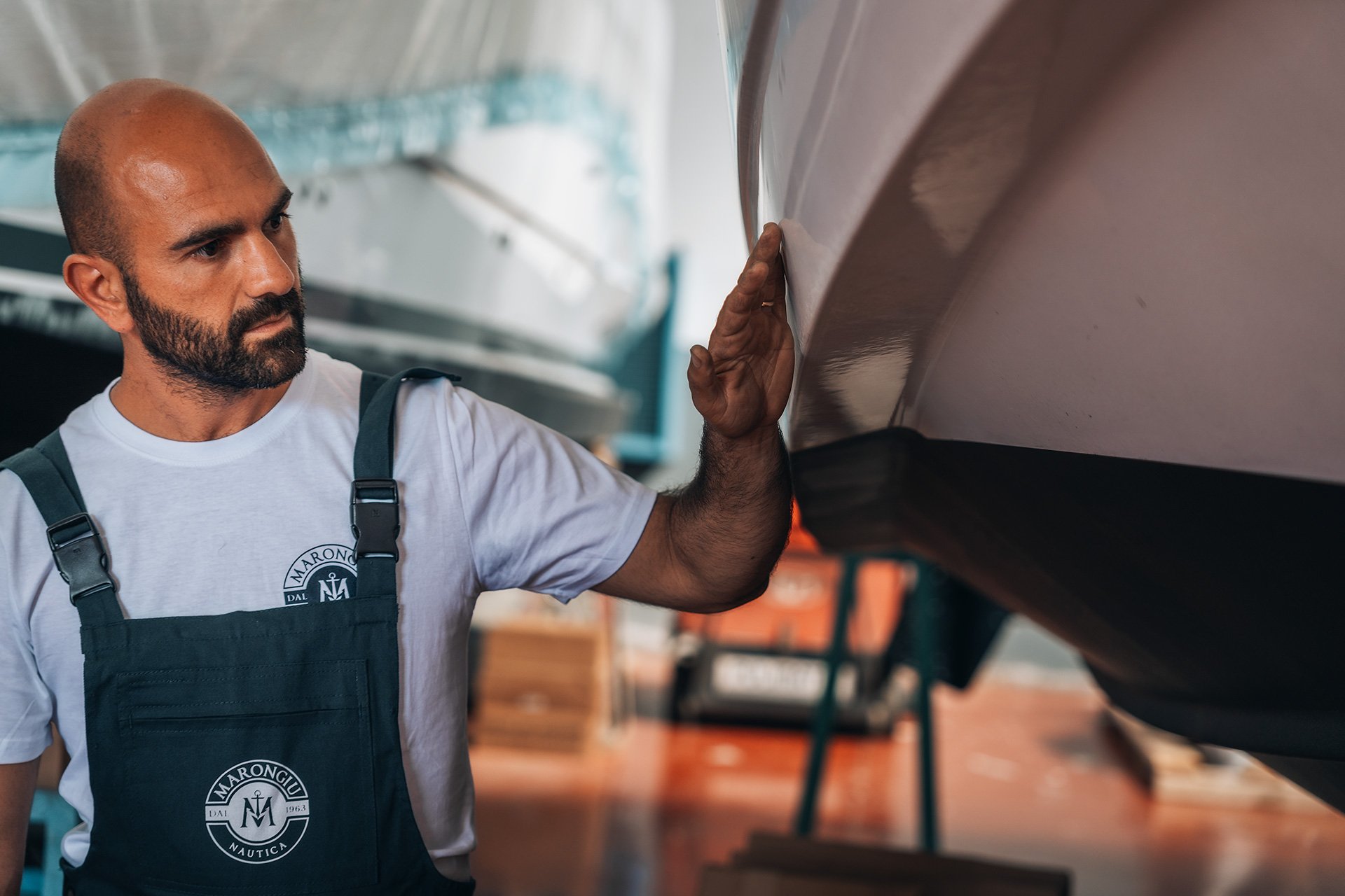Uomo con barba che controlla una barca nel magazzino, indossa una maglietta bianca e un grembiule con il logo 'Marongiu Nautica'