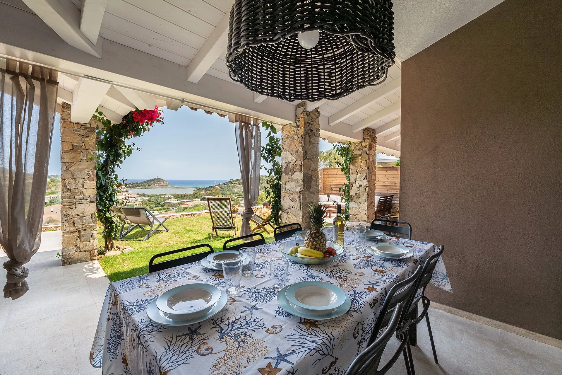 Tavola all'aperto con piatti, bicchieri e frutta, vista su giardino e mare, sotto un soffitto con lampada di vimini.