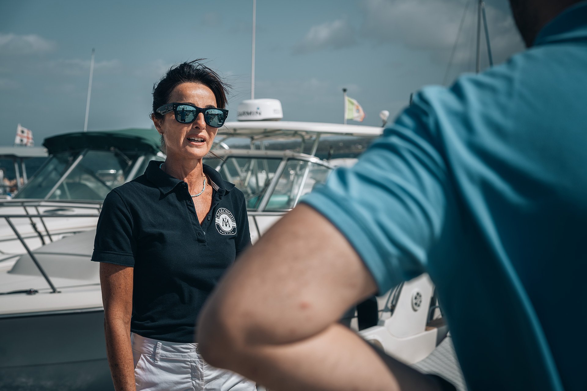 Donna in abbigliamento nautico con occhiali da sole parla con un uomo davanti a barche a Motore nel porto