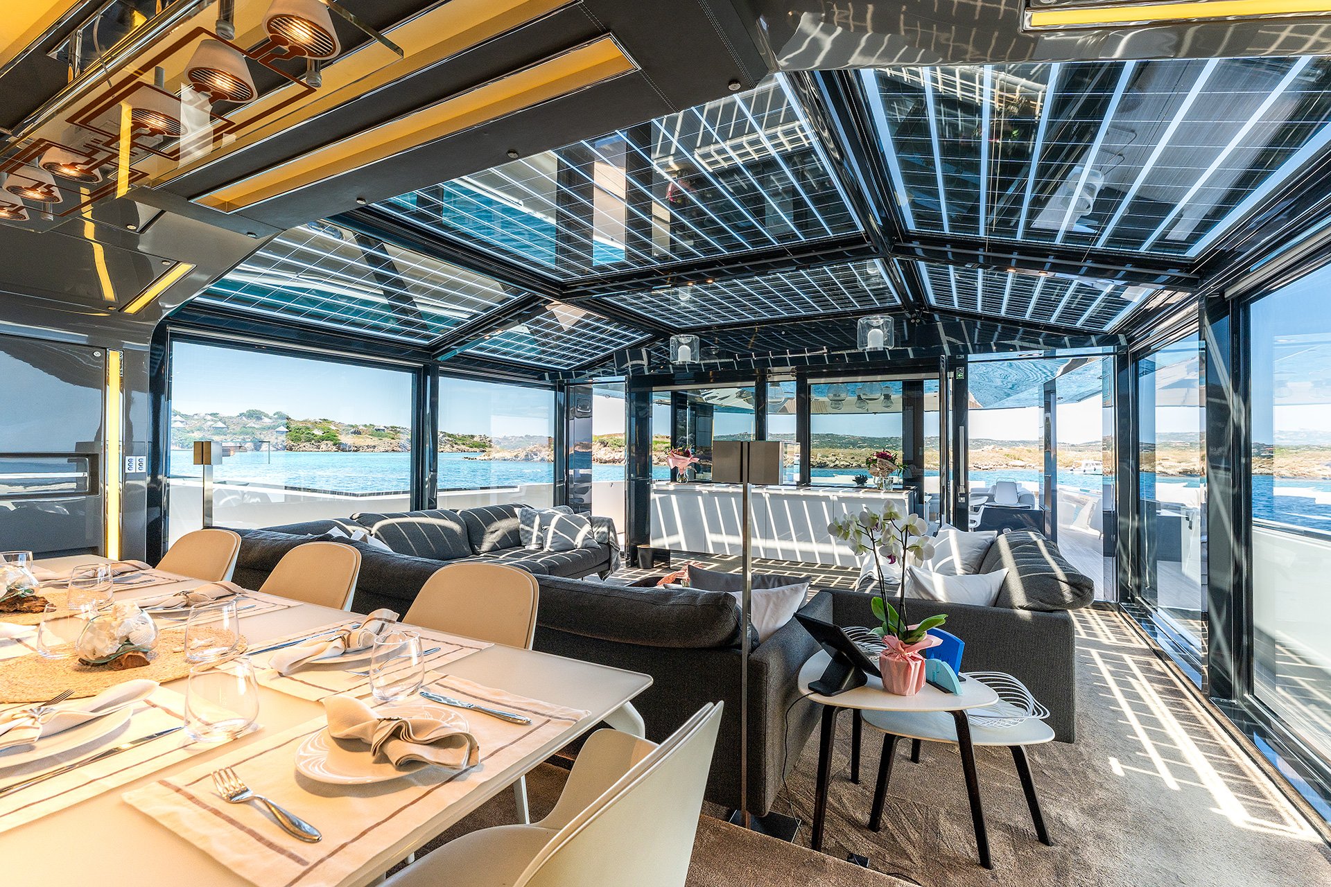 Interno di una nave con grandi finestre che mostrano un paesaggio marino e coste lontane. Arredamento moderno con divani, tavoli e fiori.