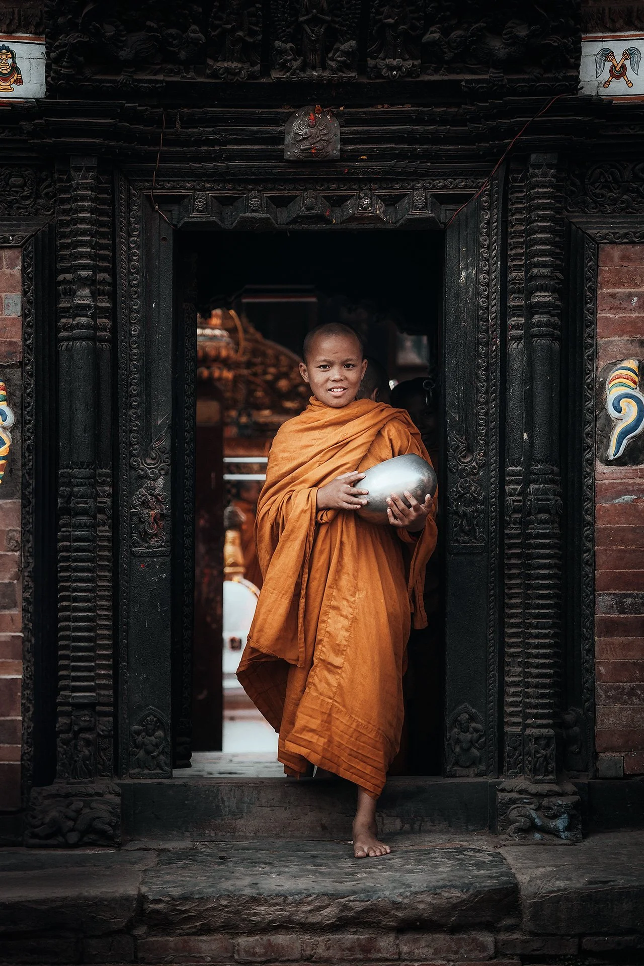 Un monaco buddhista in veste arancione si trova sulla soglia di un'abitazione tradizionale, con un vaso di metallo in mano, all'interno di un portone intagliato in legno.