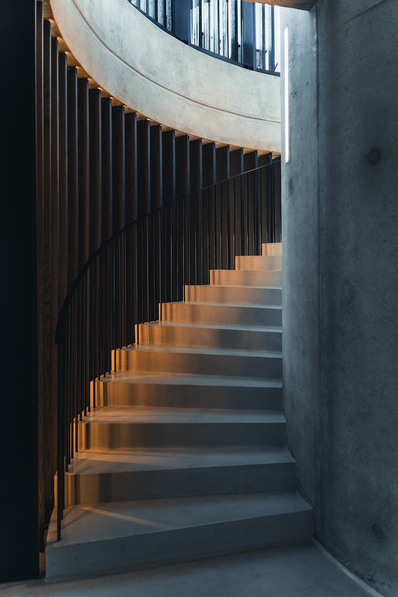 Immagine di una scala curva in cemento con ringhiera nera, illuminata da luce calda, in un'ambientazione moderna