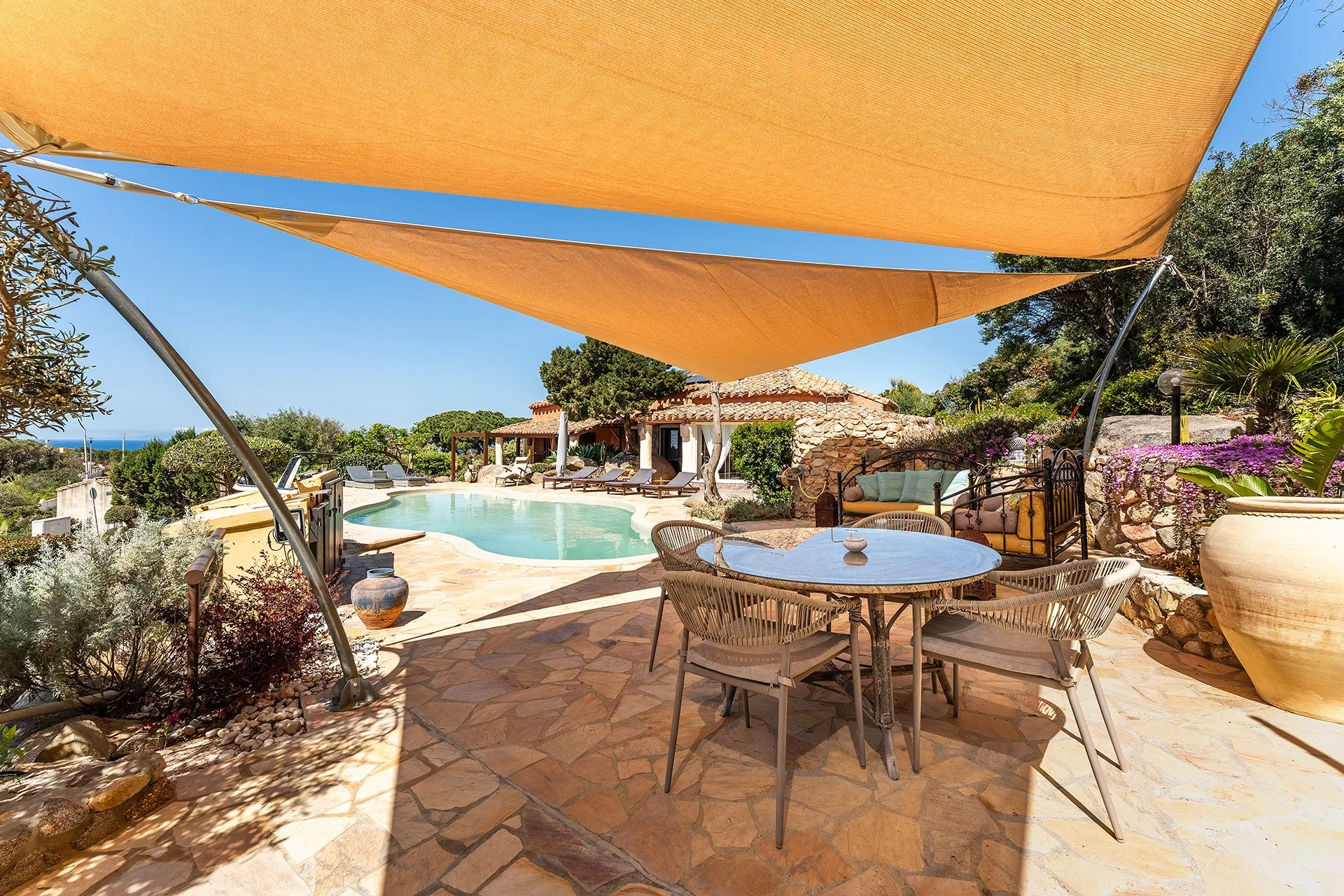 Zona relax con piscina, divani, tavolo e ampie vela parasole in un ambiente esterno colorato e pieno di verde.