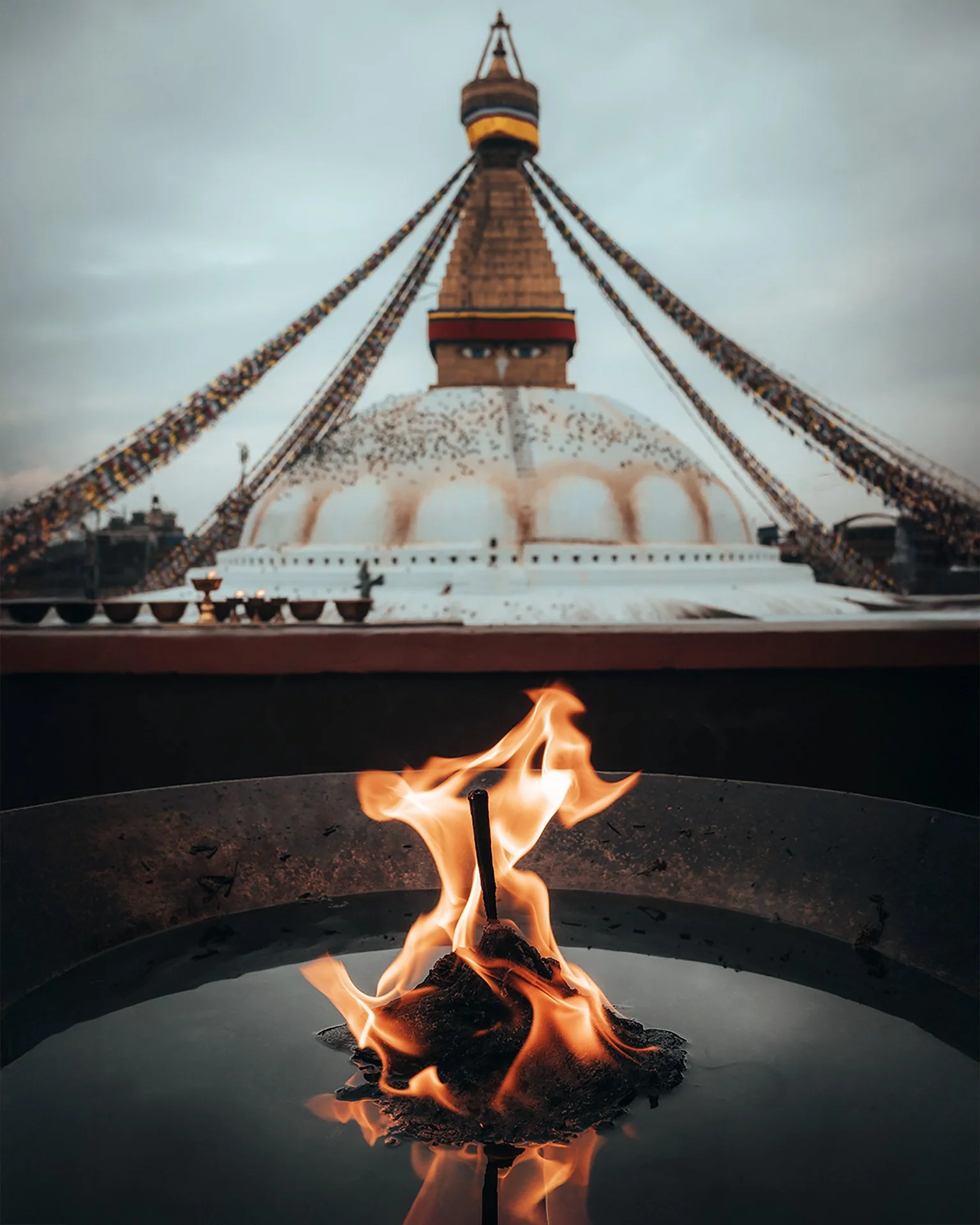 Fiamma accesa di fronte a una grande pagoda con occhi disegnati, ornata con bandiere di preghiera, sotto un cielo nuvoloso.