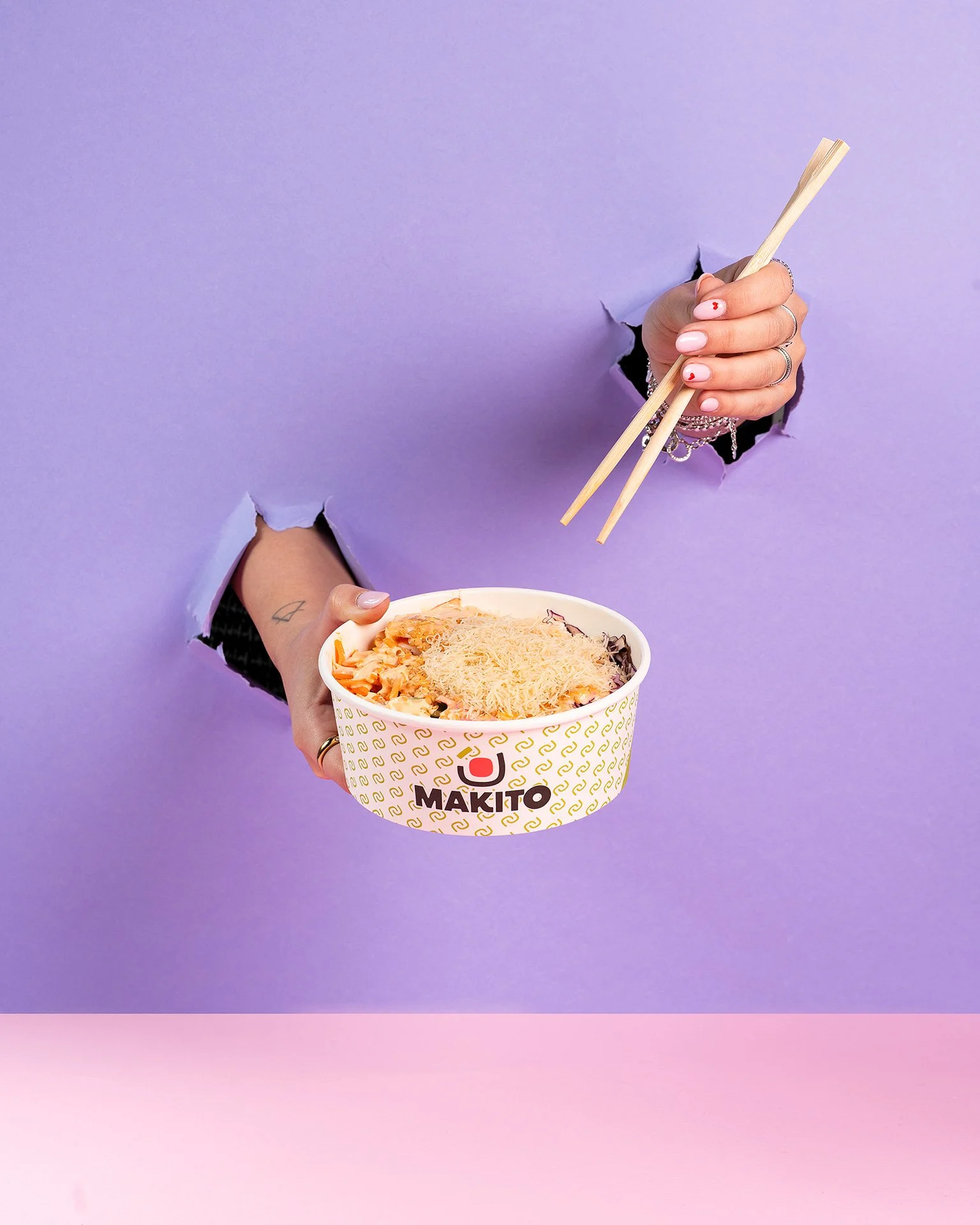 Una mano tiene un cucchiaio mentre l'altra tiene una ciotola di noodles con formaggio grattugiato. La ciotola ha il logo di Makito e il background è di colore viola con sfumature rosa.