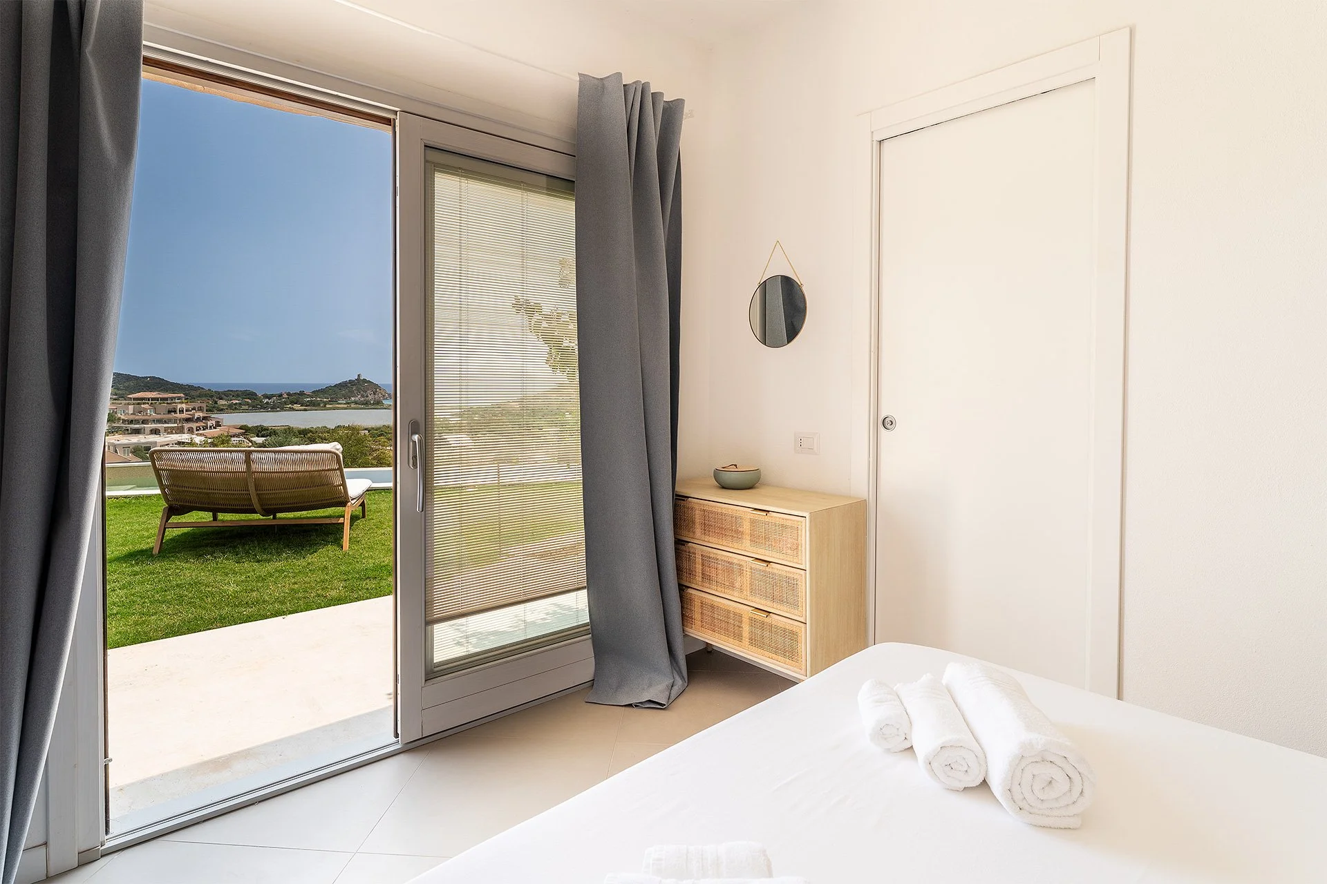 Camera da letto con porta-finestra che si apre su un terrazzo con vista mare e colline, asciugamani arrotolati sul letto, arredo semplice e divanetto di legno all'aperto scelto per rilassarsi.
