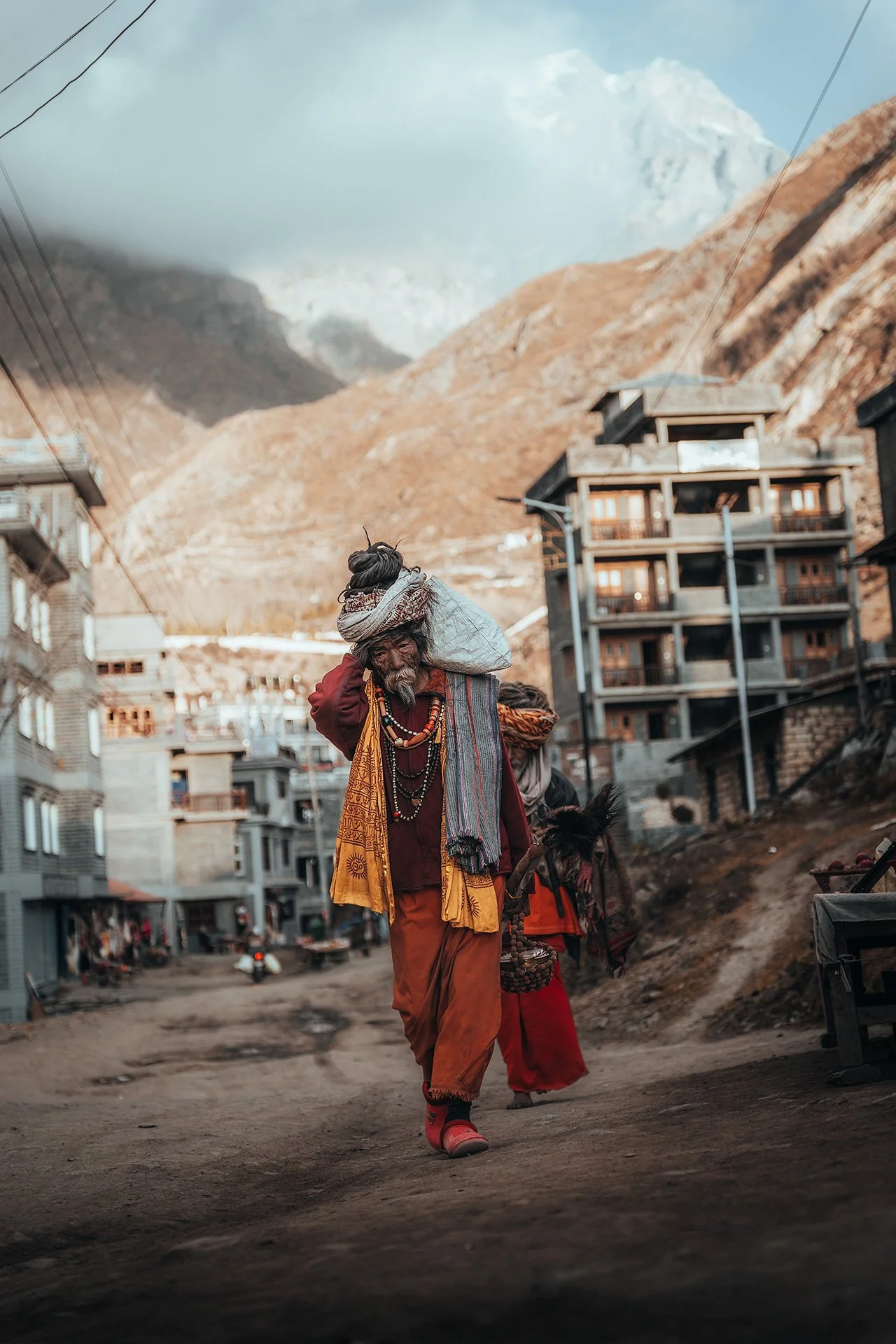 Uomo anziano vestito con abiti tradizionali, che cammina per strada a lato di altri persone e edifici, con montagne sullo sfondo.