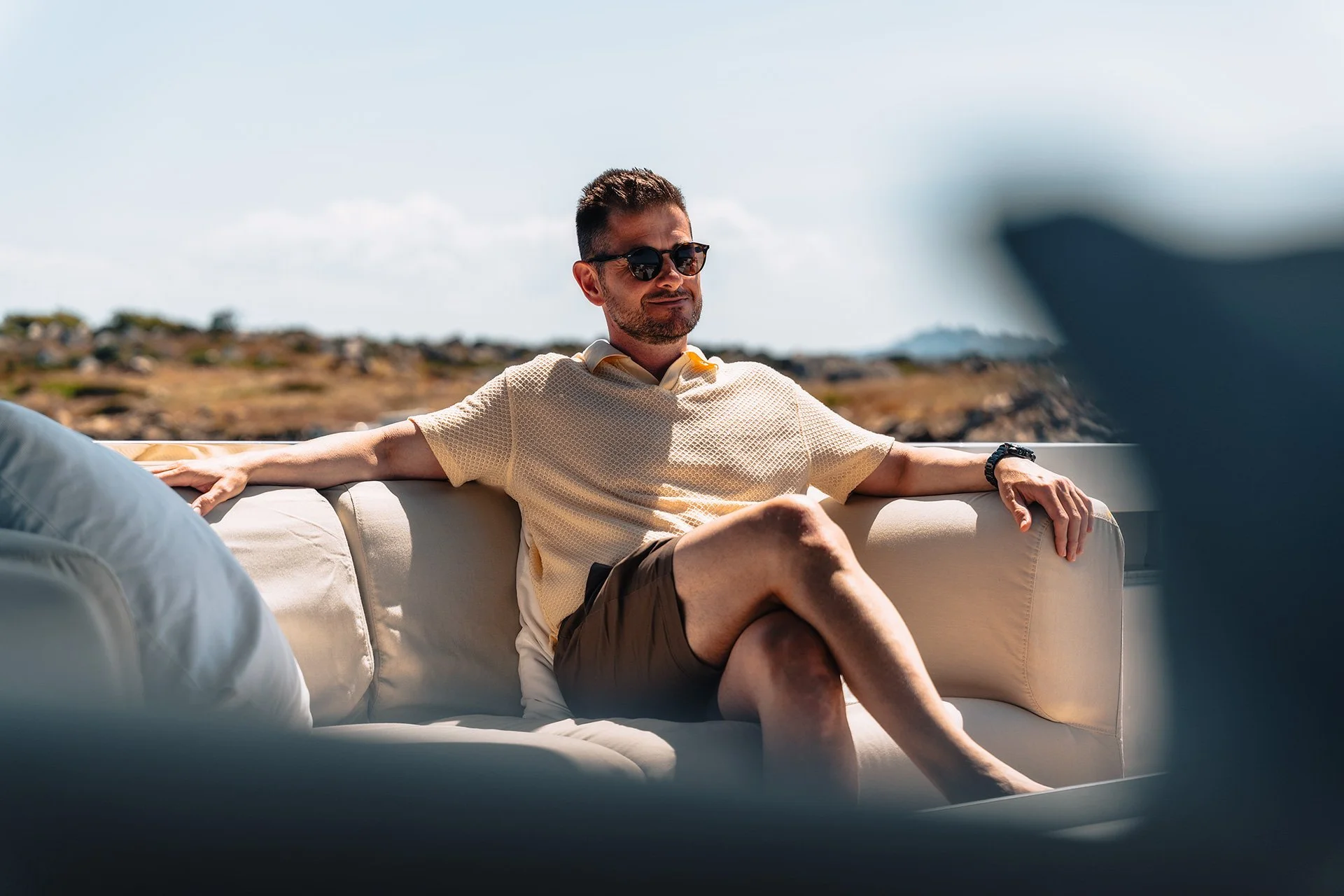 Uomo che si rilassa seduto su un divano all'aperto, indossa occhiali da sole e abbigliamento estivo, con un paesaggio di scogliera e cielo azzurro sullo sfondo.
