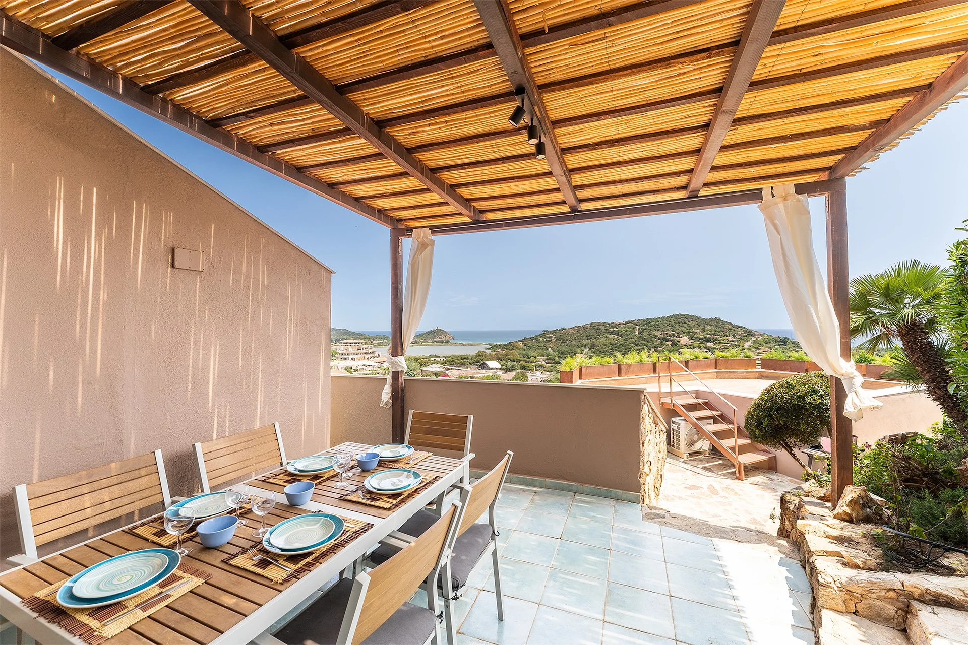 Terrazza con tavolo da pranzo e piatti, vista sul mare e colline, tettoia in bambù, piante verdi