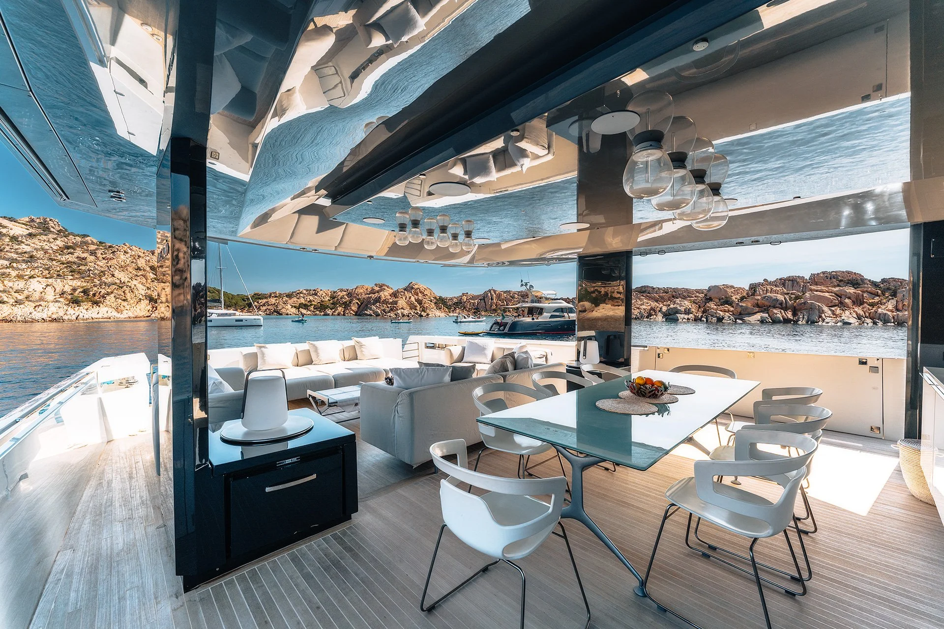 Interno di una casa sul mare con vista panoramica su barche e rocce, arredamento moderno con divani bianchi, tavolo da pranzo con sedie bianche, lampade sospese in vetro e cucina con piano nero.