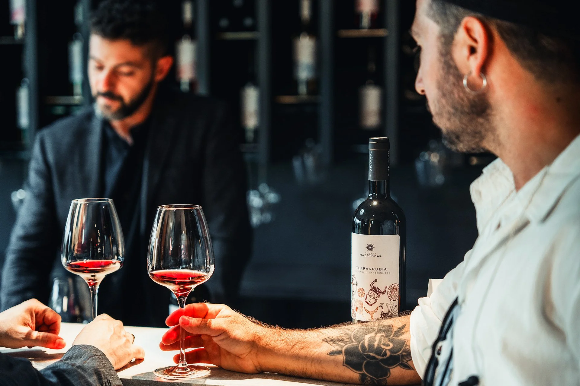Due persone stanno brindando con due bicchieri di vino rosso in un ristorante. Sul tavolo c'è una bottiglia di vino, e in background si vedono scaffali con bottiglie.