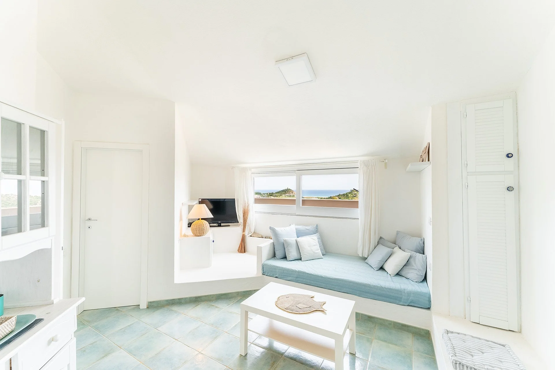 Interno di una camera da letto con pareti bianche, letto con cuscini e una finestra che mostra un paesaggio con vista mare.