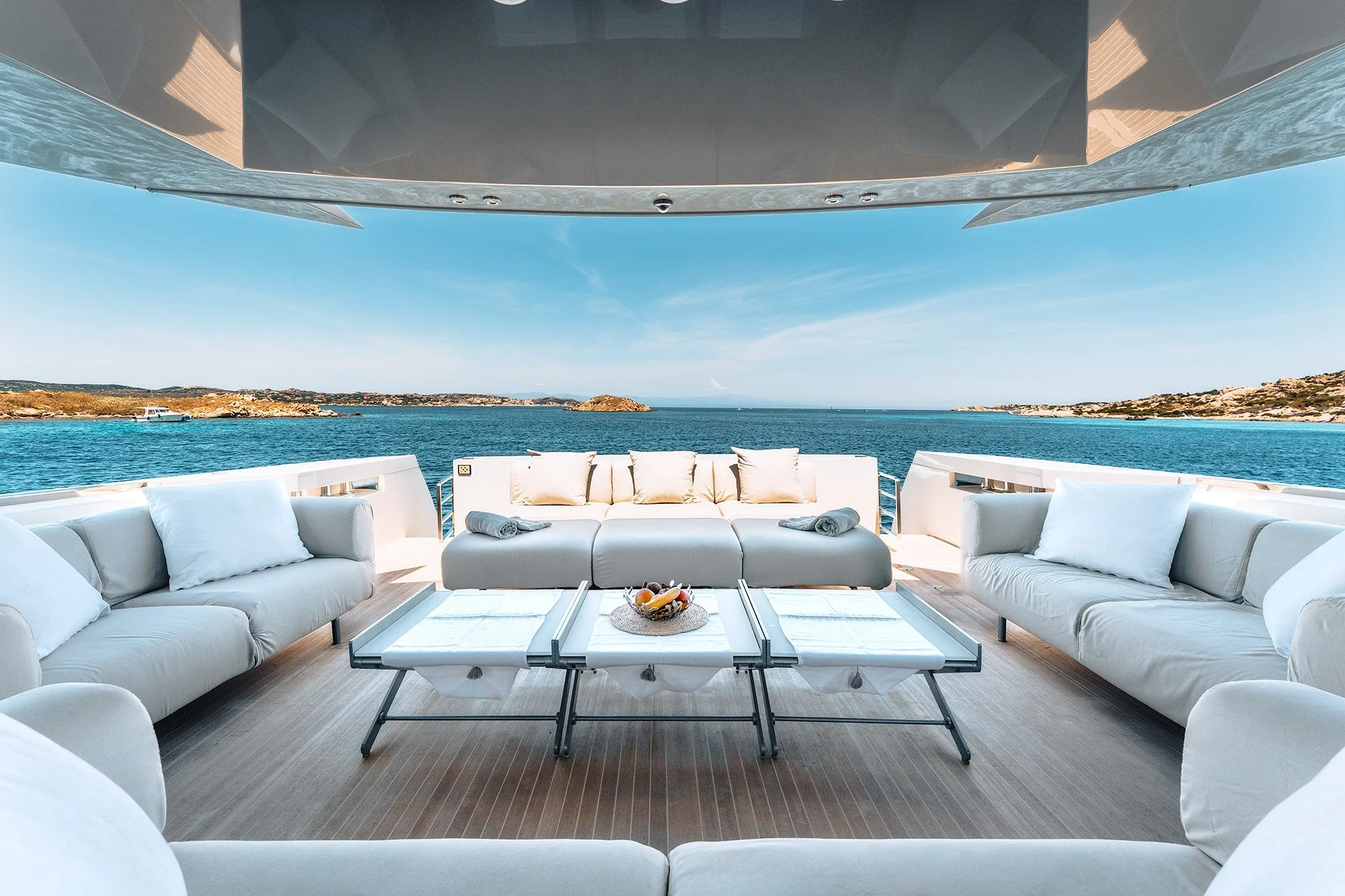 L'immagine mostra una zona lounge di un yacht con divani bianchi e cuscini, tavolino centrale con frutta, vista sul mare e coste sullo sfondo, sotto un grande parapetto e sopra una copertura in metallo lucido.