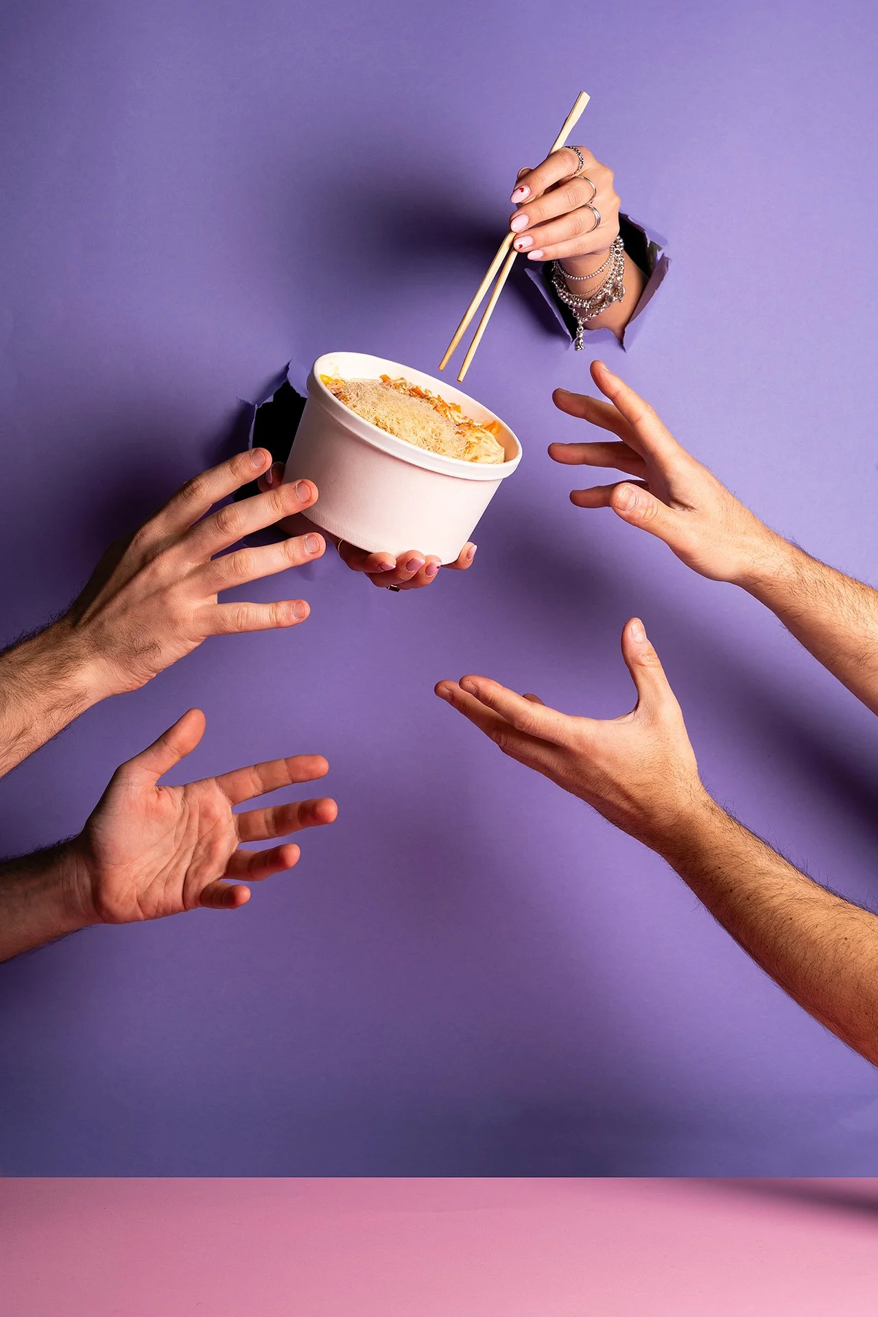 Mani di diverse persone che tengono o offrono una ciotola di ramen con chopsticks contro uno sfondo a due colori, lavanda e rosa.