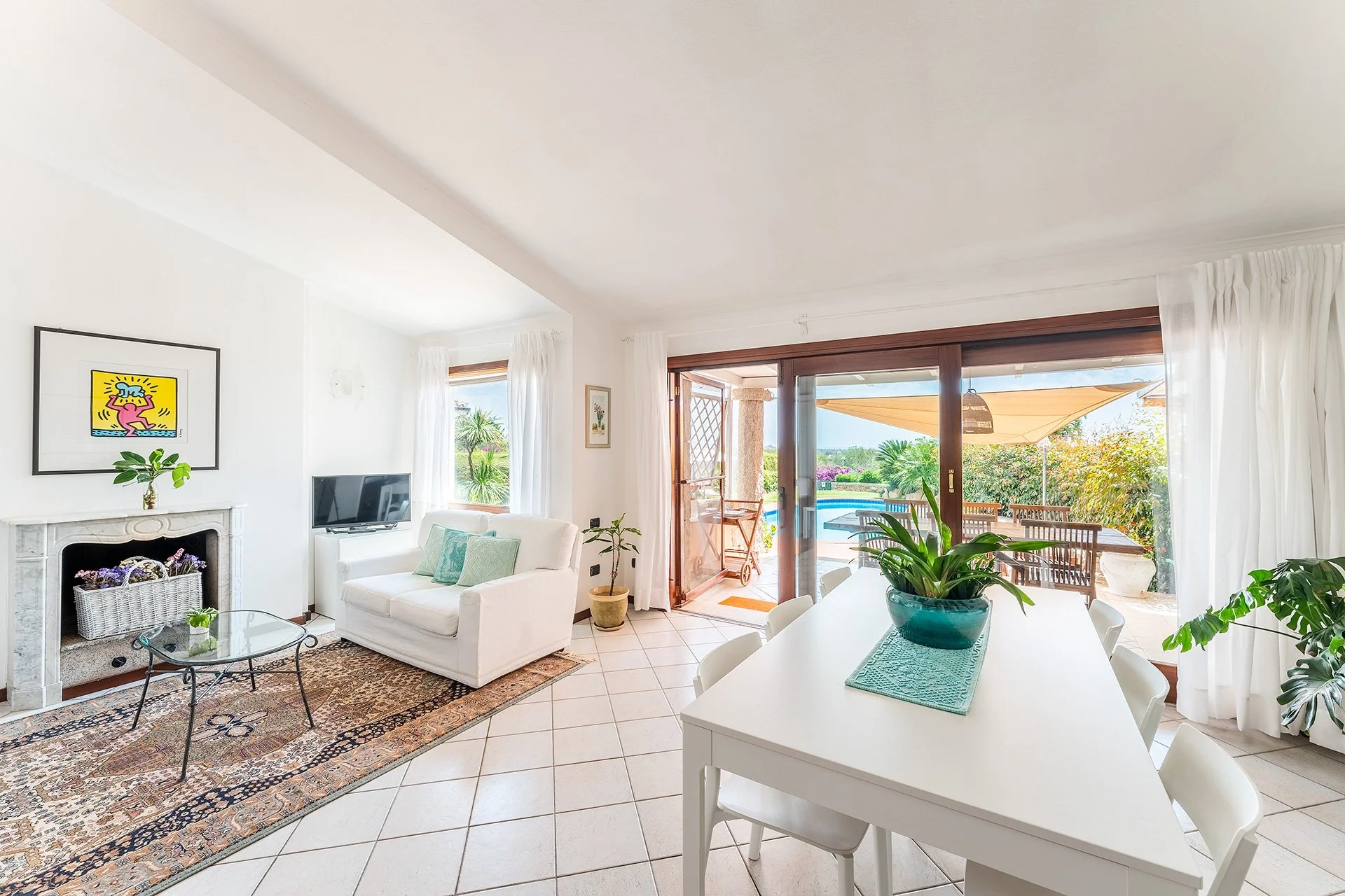 Interno di una sala soleggiata con pareti bianche, camino decorato, divano bianco con cuscini, tavolo tondo di vetro, piante in vaso, grande finestra con tende bianche, vista verso un patio con tavolo e sedie e piscina nel giardino